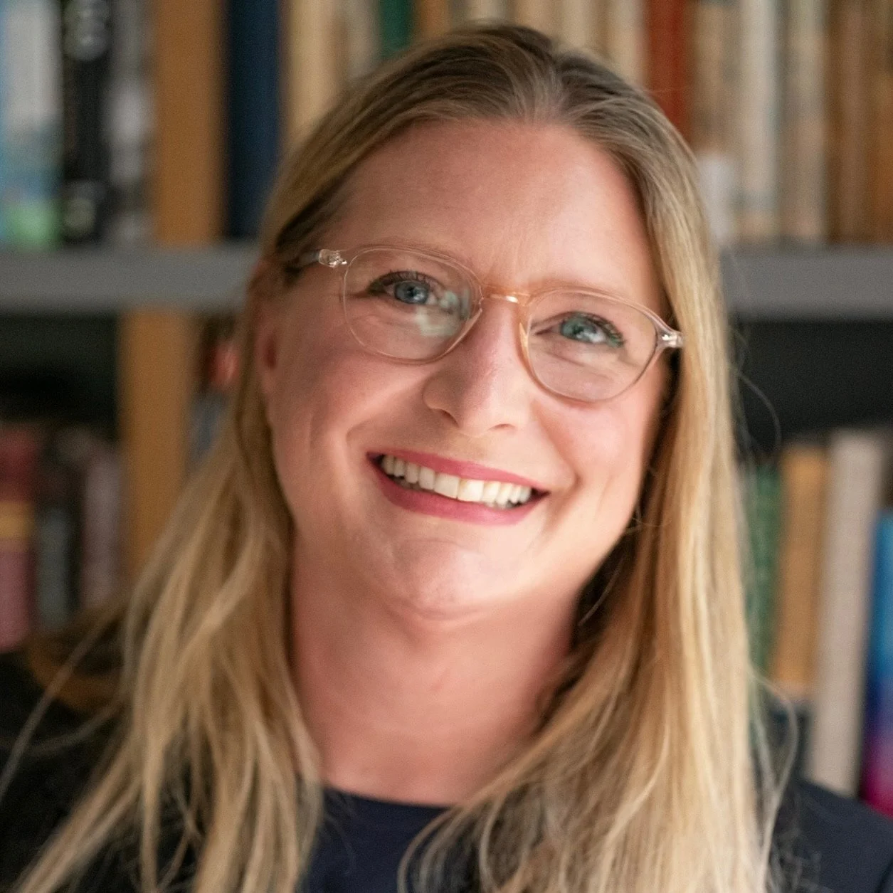 A woman with long blonde hair wearing glasses smiling in front of a bookshelf.