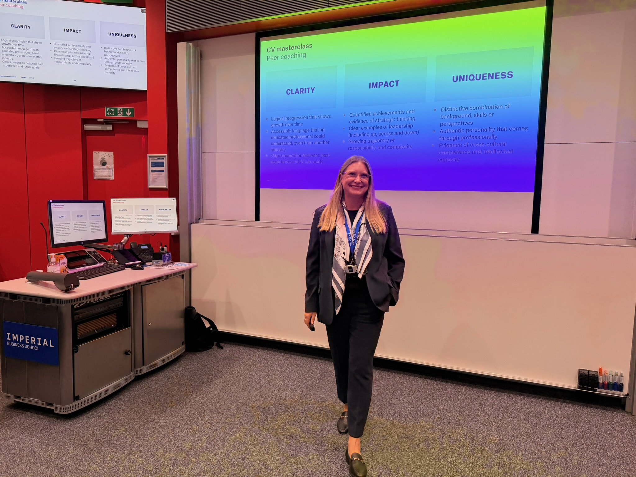 Woman in business attire walking in front of a presentation screen with slides about CV masterclass and peer coaching at Imperial Business School, conference room setting with technical equipment on the table.