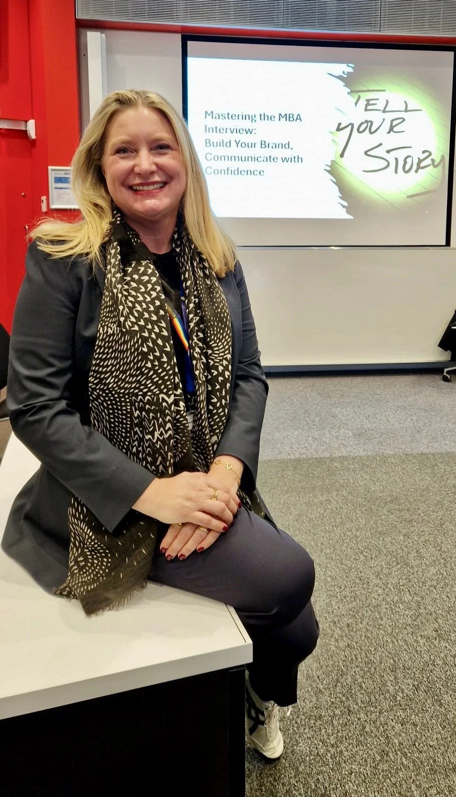 A woman sitting on a white table in a conference room, smiling at the camera. Behind her is a presentation screen with a slide titled 'Mastering the MBA Interview: Build Your Brand, Communicate with Confidence' and the phrase 'Tell Your Story' in green handwriting.