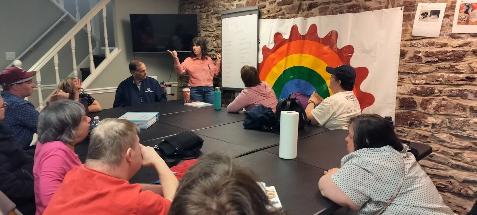 Group meeting with a woman speaking in front of a board, rainbow banner in background, attendees sitting at a table.