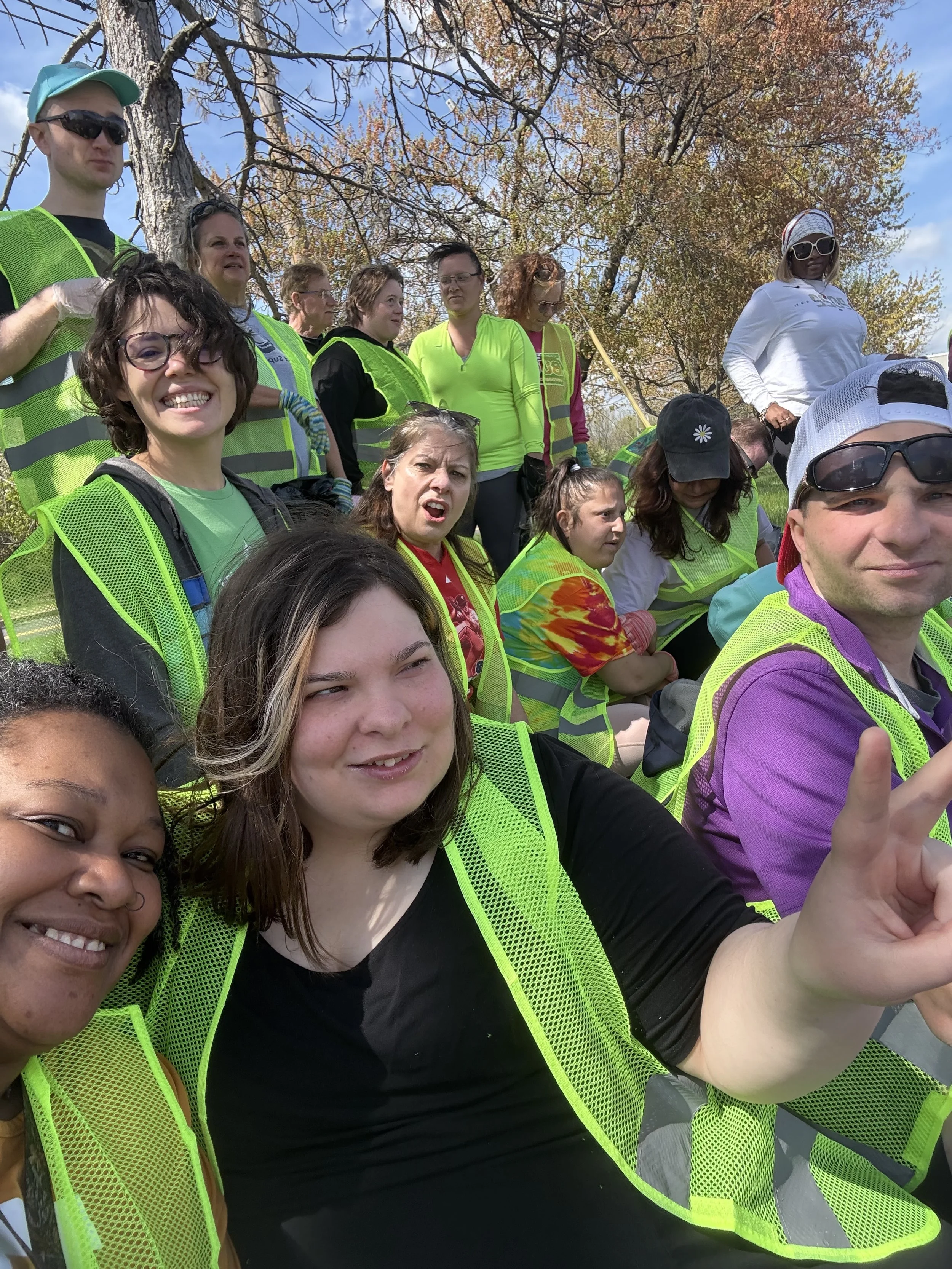 A group of people wearing bright yellow safety vests outdoors, some are taking a selfie, with trees and a blue sky in the background.