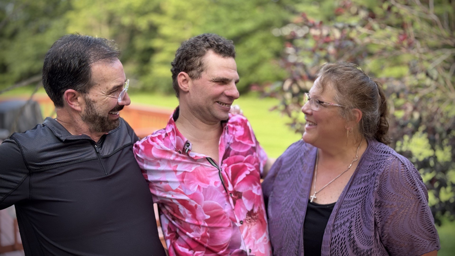 Three people standing close together outdoors, smiling and talking with each other, with trees and greenery in the background.