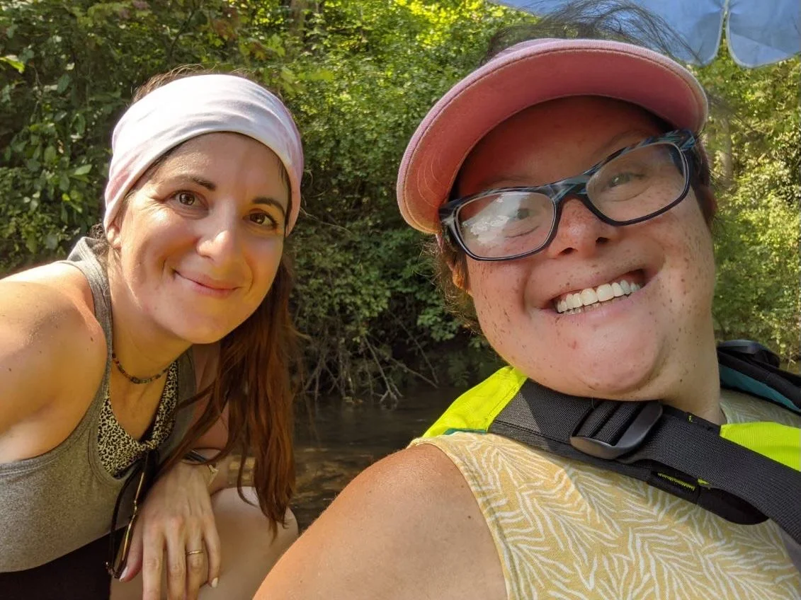 Two smiling women outdoors near a river, surrounded by green trees, taking a selfie.