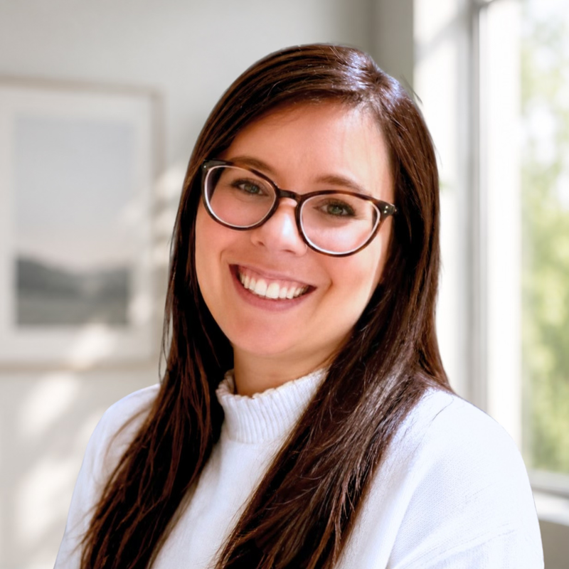 Smiling woman with long brown hair and glasses, wearing a white top, indoors near a window with a blurred background.