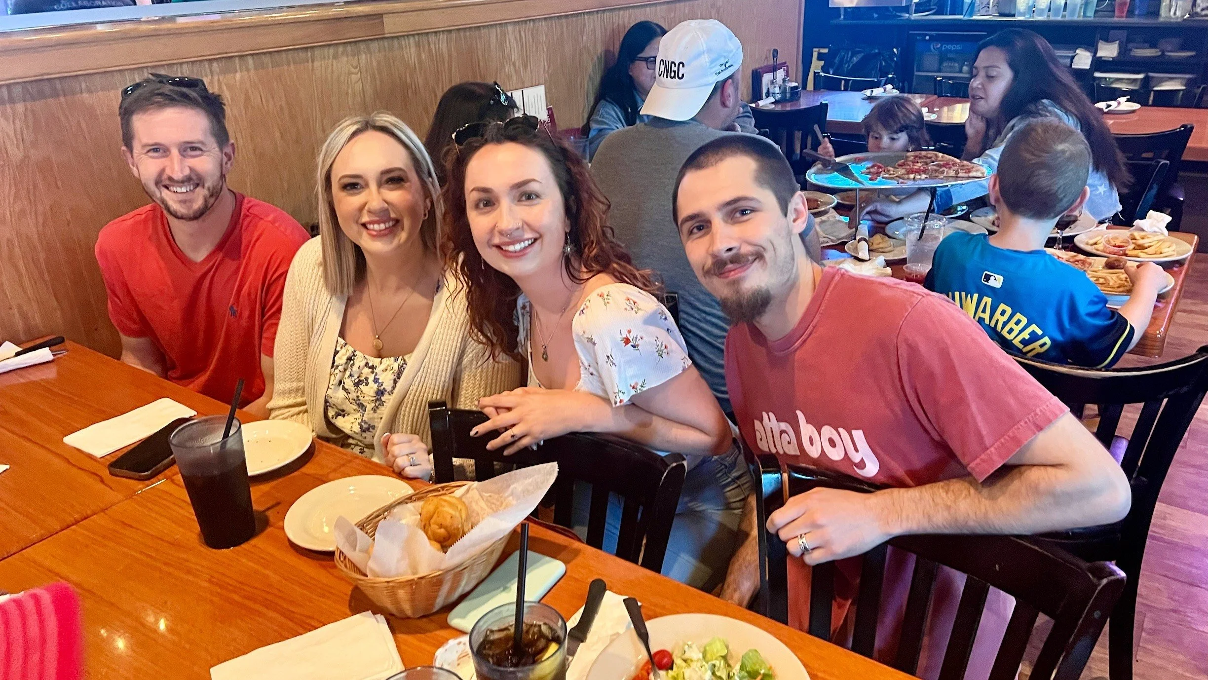 A group of four adults sitting at a restaurant table, smiling at the camera, with two children and pizza in the background.