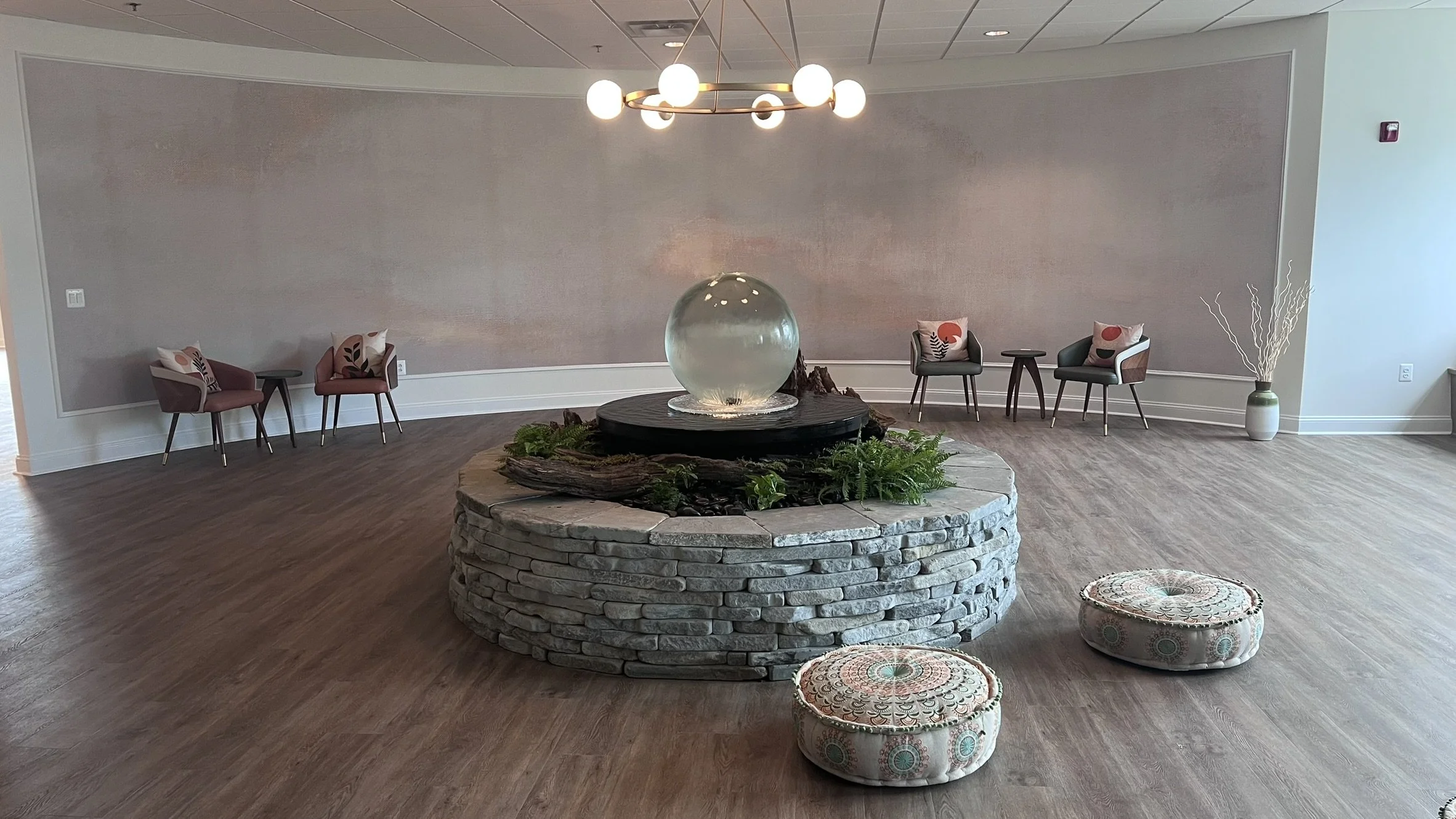 Interior of a waiting area with a central water fountain, small chairs with decorative pillows along the wall, and two patterned cushions on the floor, with a modern chandelier overhead.