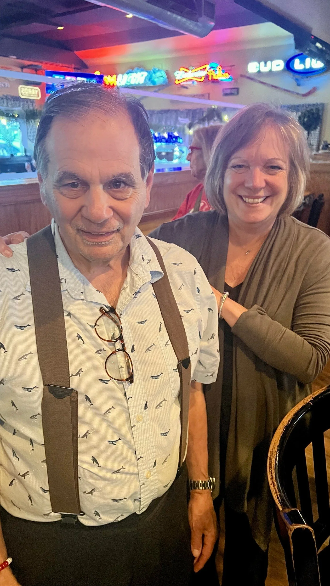 A man and woman smiling in a bar with neon signs on the wall behind them.