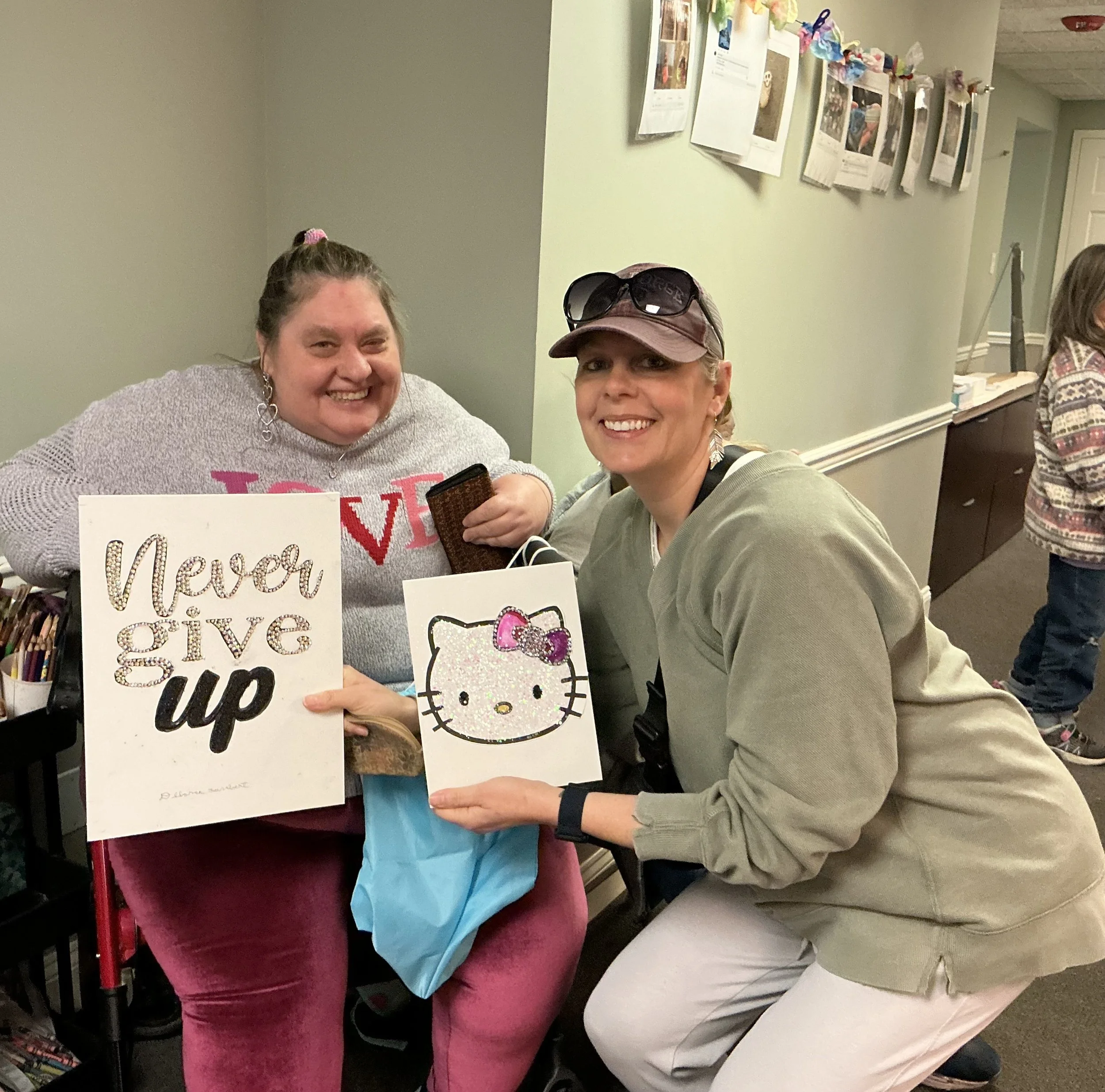 Two women smiling indoors, one holding a "Never give up" sign, the other holding a Hello Kitty gift bag.