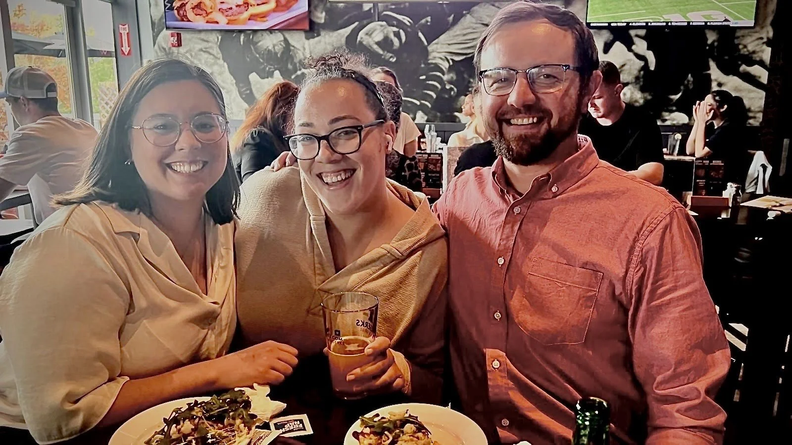 Three people smiling at a restaurant, with plates of food and drinks in front of them, and other patrons dining in the background.