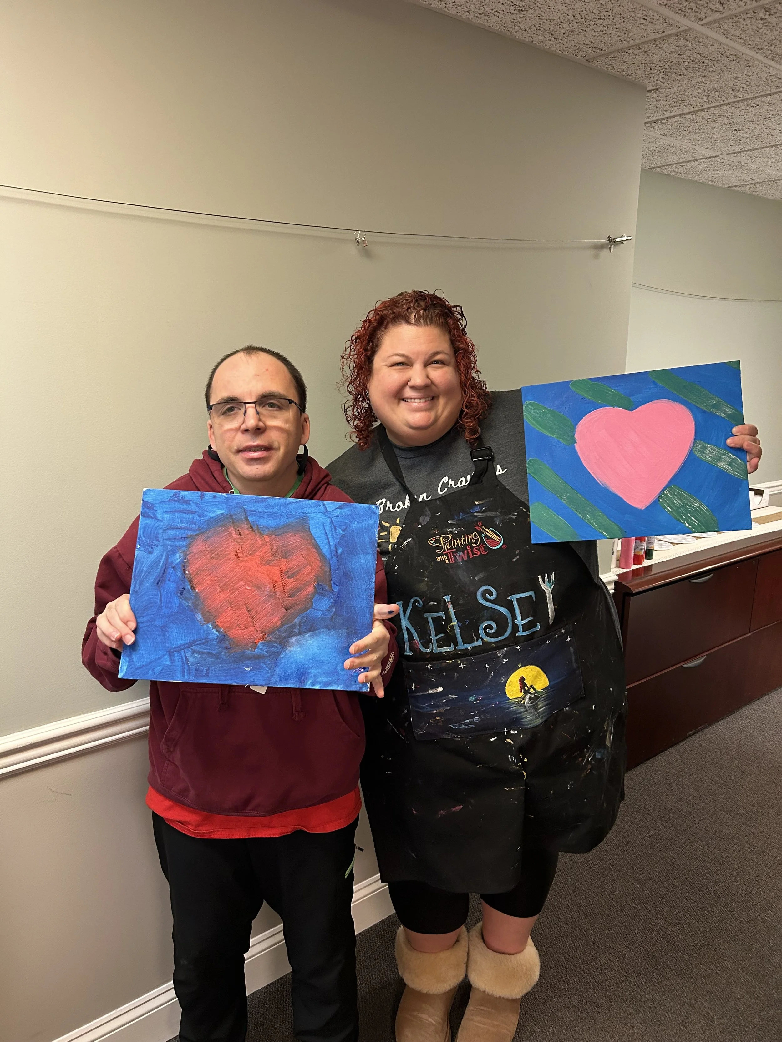 A man and a woman standing indoors, smiling, each holding a painting of a heart. The man's painting features a red heart on a blue background, and the woman's painting features a pink heart with green leaves on a blue background. The woman is wearing a black apron with painted details and the name 'Kelsey' on it. They are in front of a light-colored wall and a wooden counter.