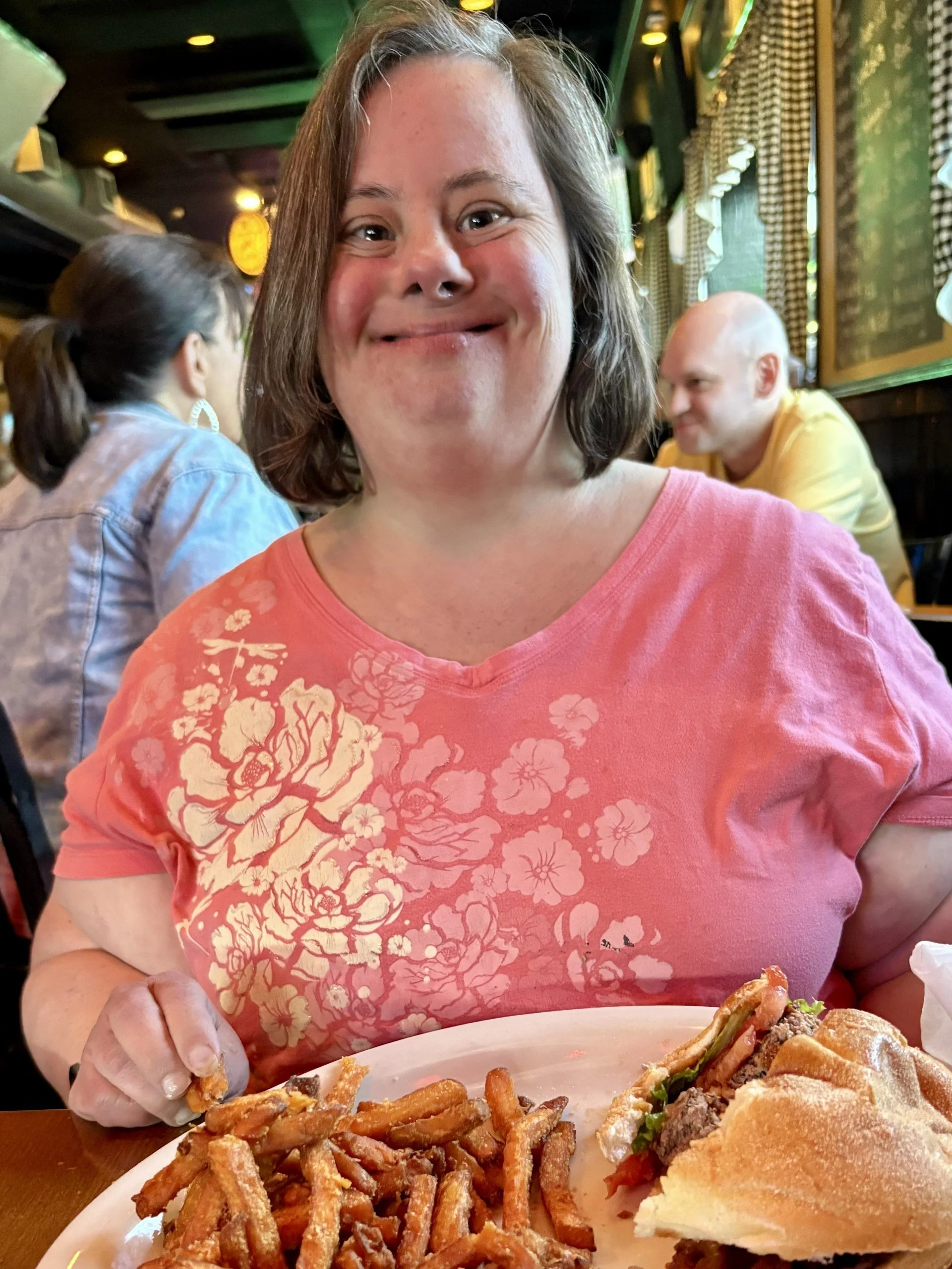 A woman with short brown hair wearing a pink floral t-shirt, smiling at the camera, sitting in a restaurant with a plate of fries and a sandwich in front of her.