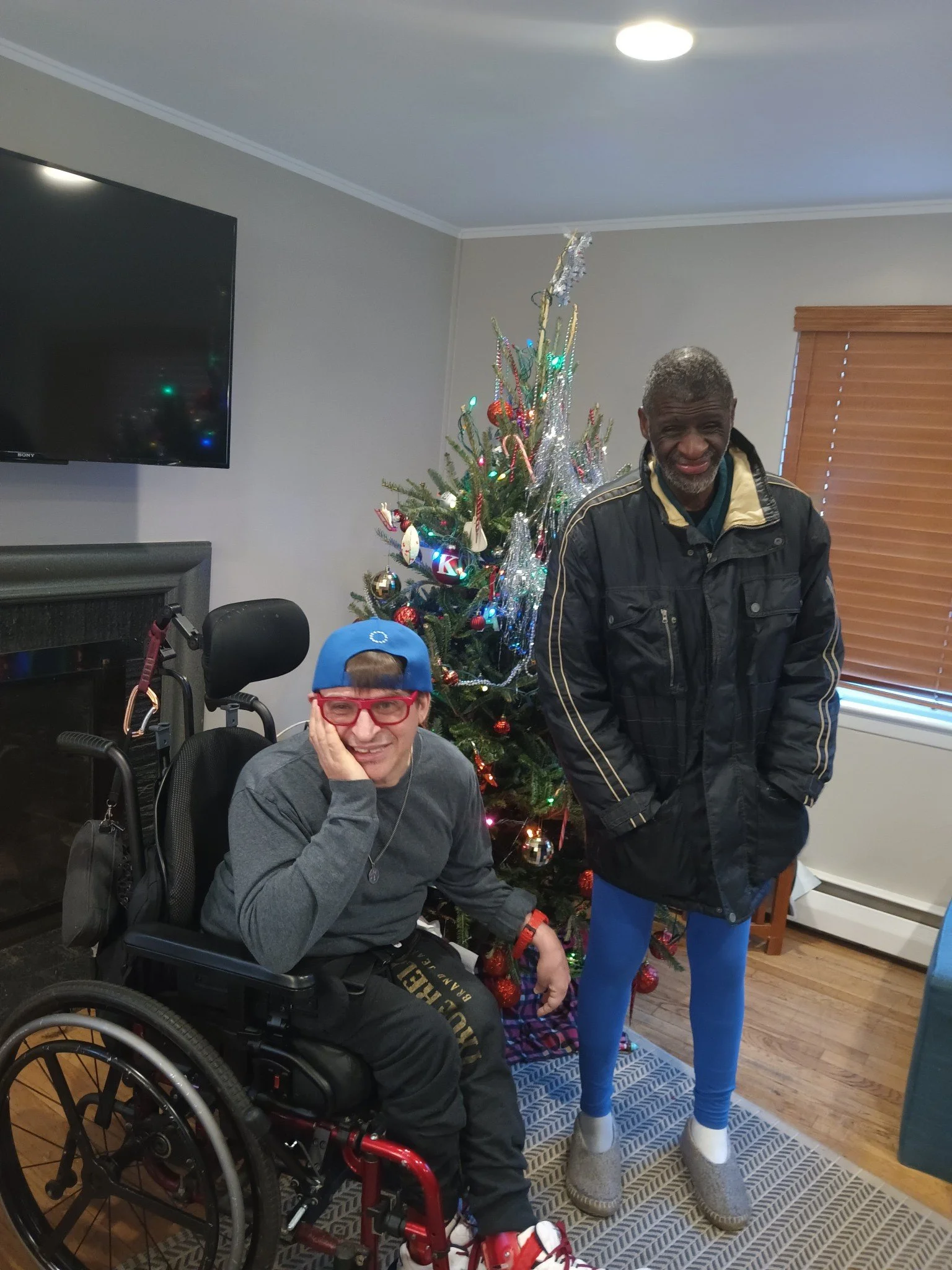 Two people in a room with a decorated Christmas tree. One person is sitting in a wheelchair wearing red glasses and a blue cap, and the other person is standing next to them wearing a dark jacket. There is a television mounted on the wall and a window with blinds.