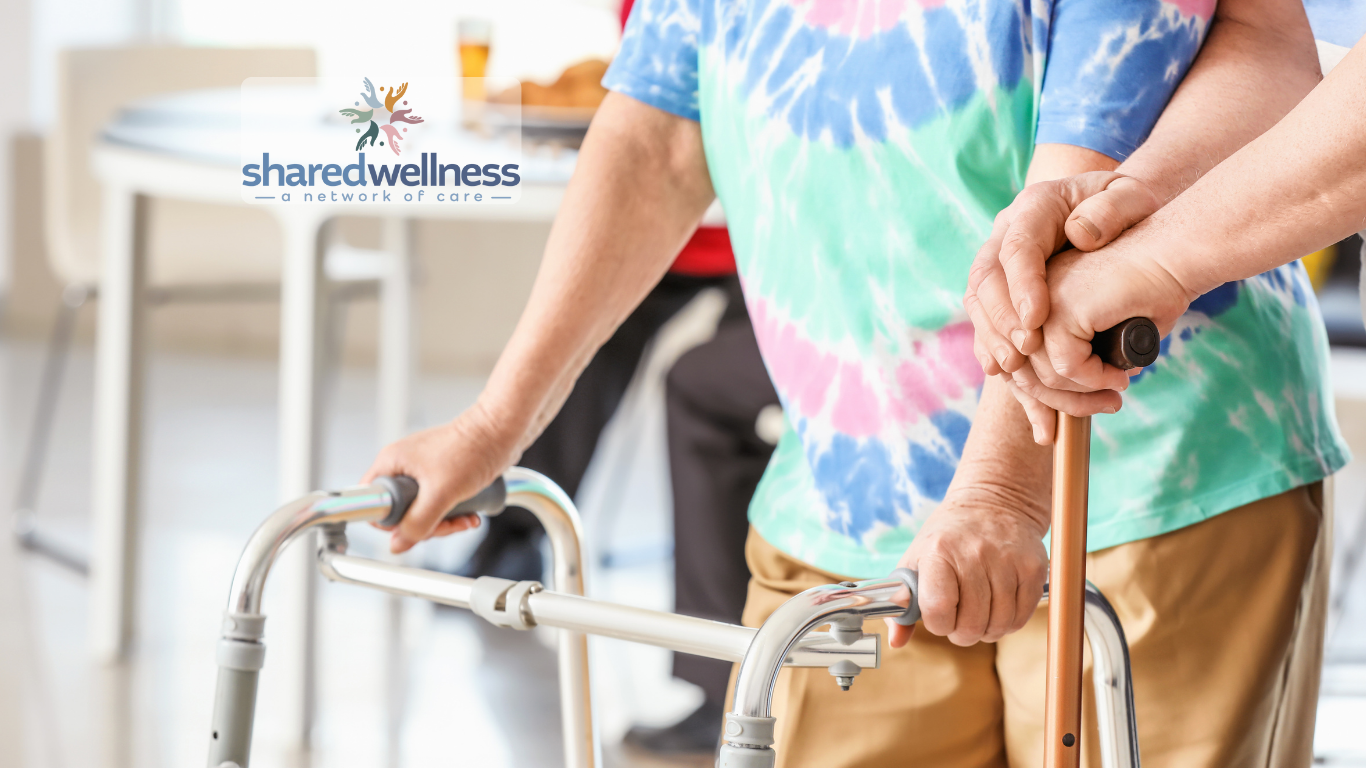 A young man in a motorized wheelchair with head and neck support, making a playful face with his tongue out. A woman with glasses, wearing a patterned pink and white shirt, is standing beside him, smiling. The background shows a door, a Philadelphia Eagles banner, and framed photos on the wall.