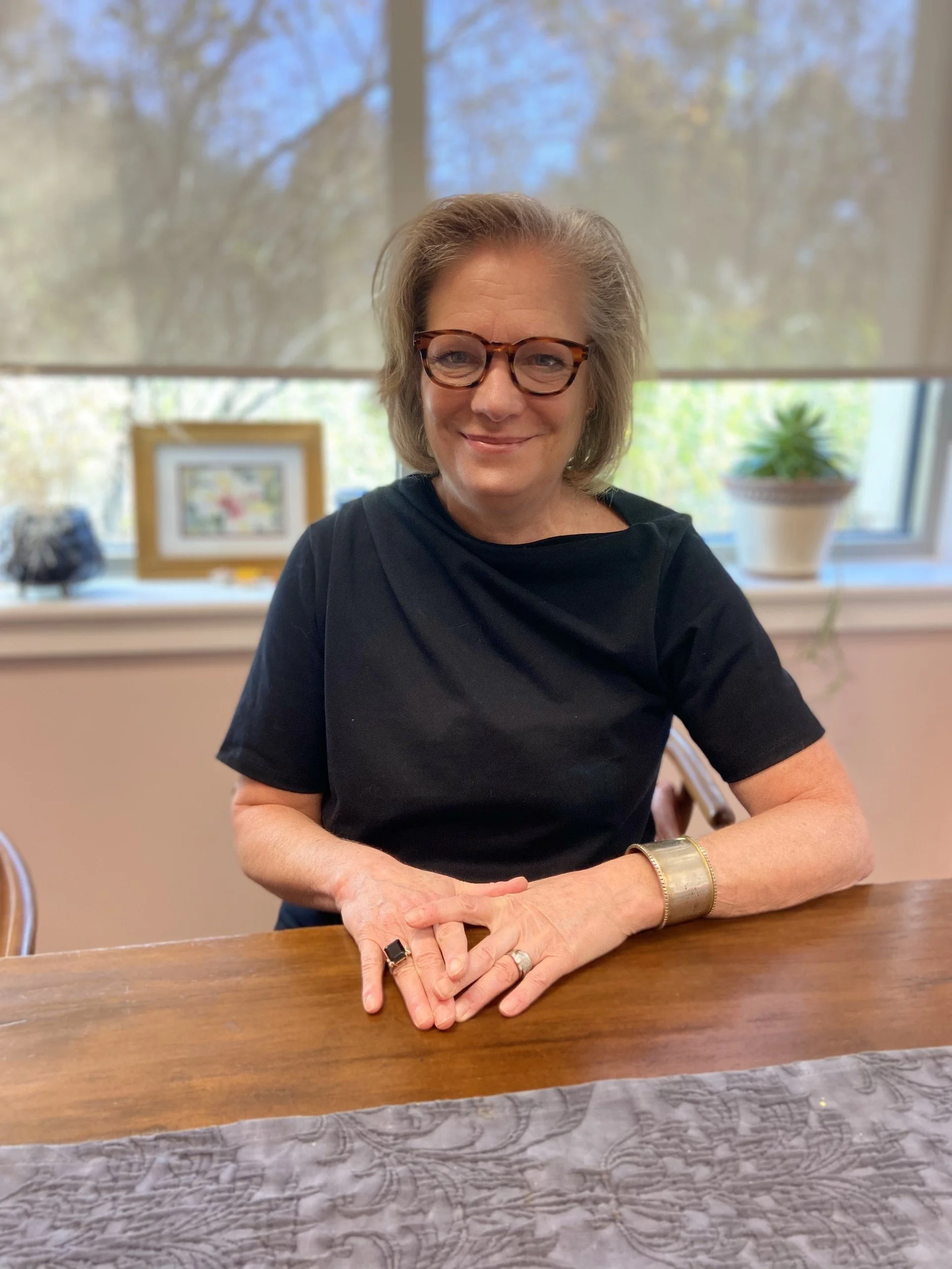 Woman wearing glasses and black top sitting at a wooden table with a textile, framed picture, and potted plant in the background.