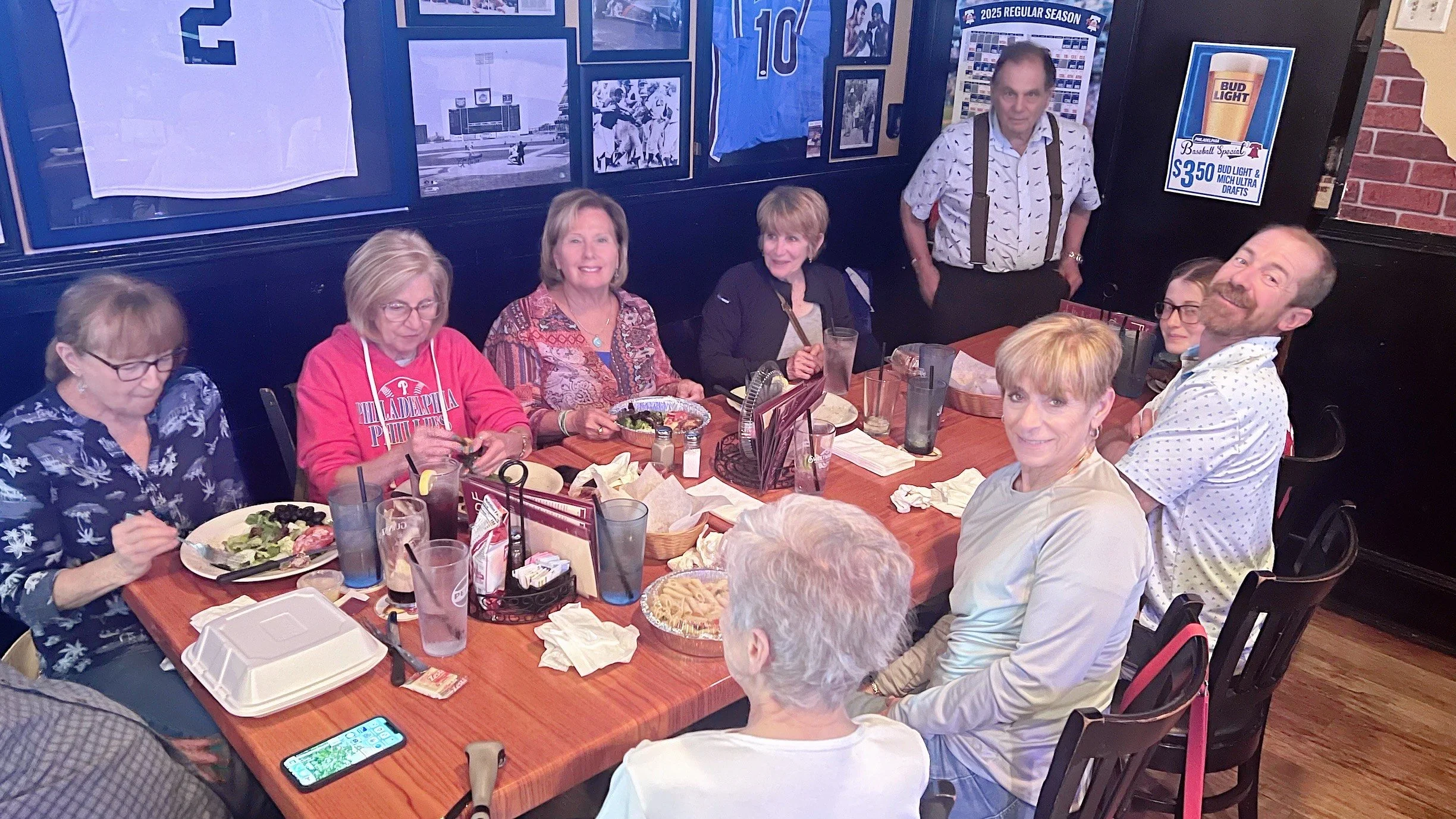 A group of nine people, mostly women, are gathered around a long wooden table at a restaurant, some eating and some smiling at the camera. The table has plates of food, drinks, and condiments, with framed sports memorabilia and photos on the wall behind them.