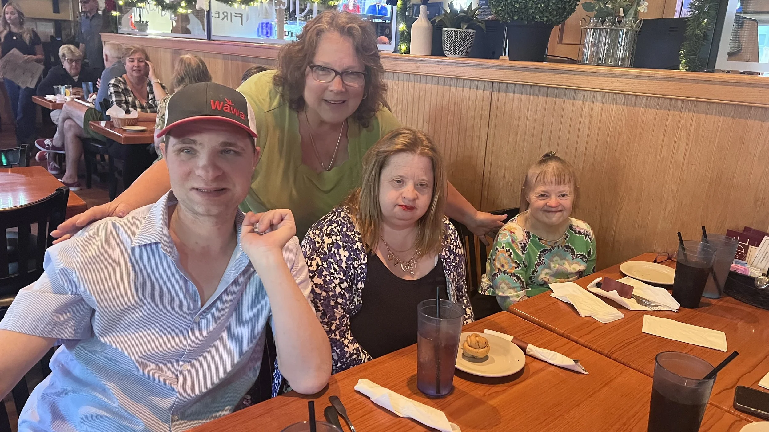 Four women and one man sitting at a restaurant table, with some women standing behind. The table has plates, cups, napkins, and utensils. The background shows other diners and decor with potted plants.