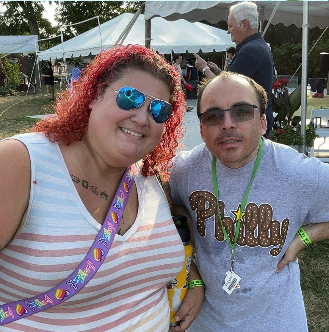 Two people smiling outdoors at a festival or outdoor event, with tents and other attendees in the background.