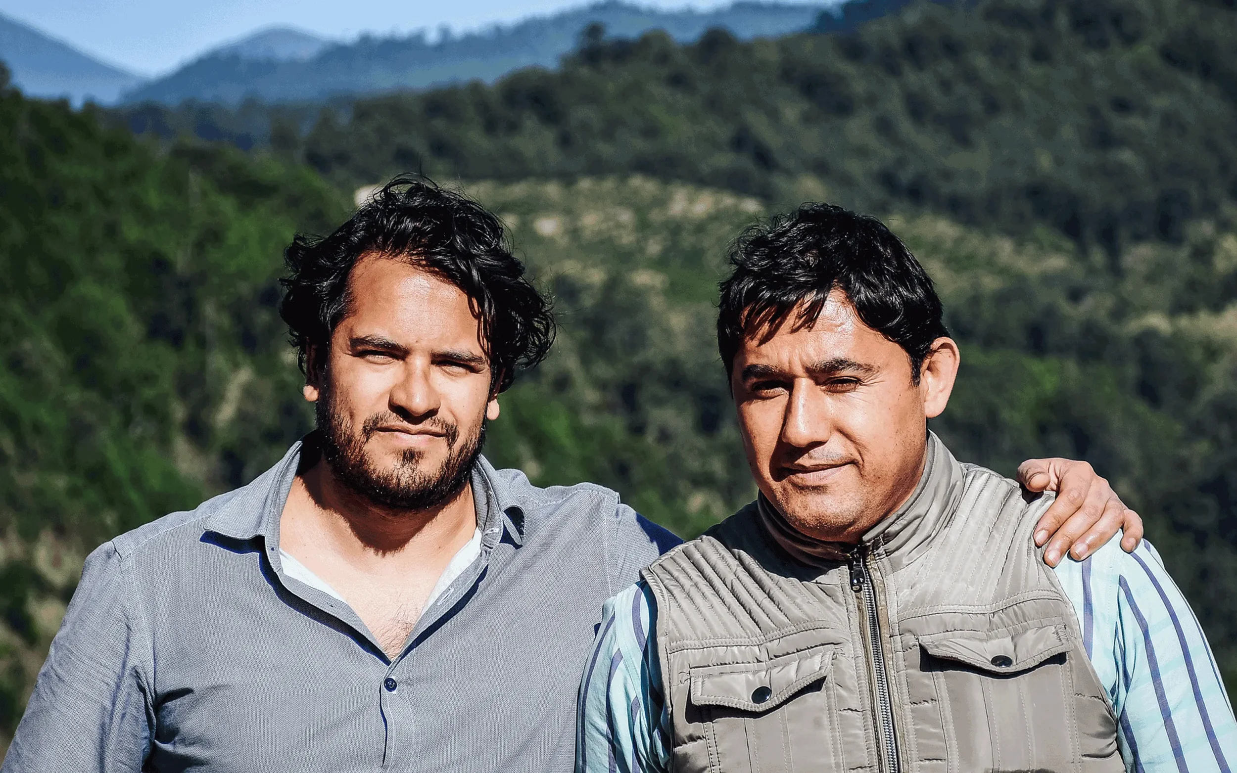 Two men standing outdoors in front of lush green hills avocado farm, one has his arm around the other's shoulder.