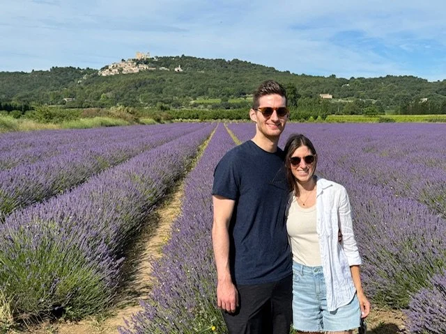 A man and woman standing together in a lavender field with a castle on a hill in the background.