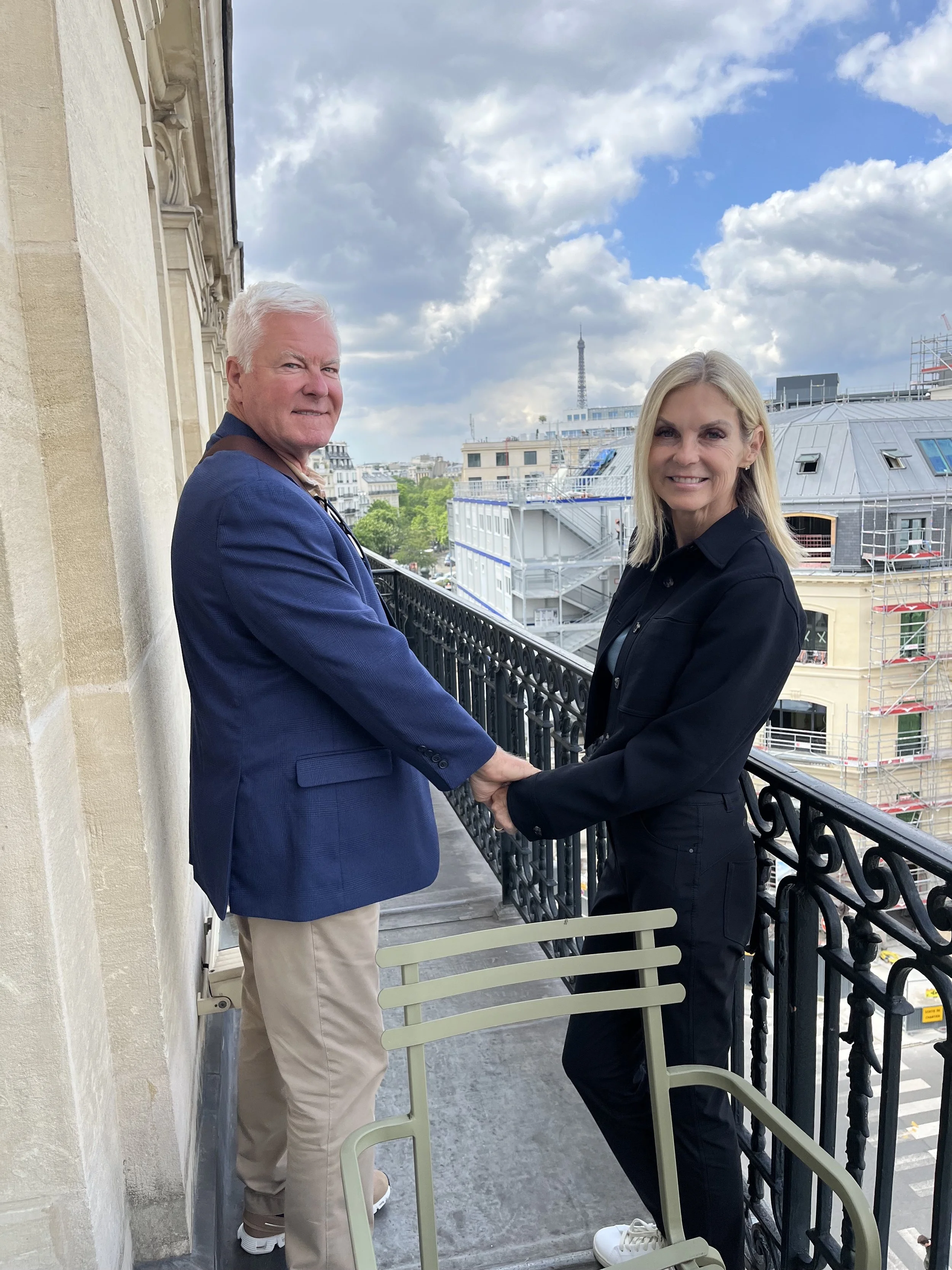 A man and a woman hold hands on a balcony with Paris skyline in the background, including the Eiffel Tower and cloudy sky.