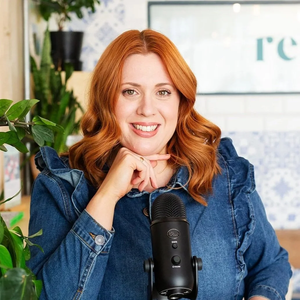 Bethany Helliwell-Smith, Charity Coaching, founder of Awen Coaching, with red hair, wearing a floral blouse, sitting at a wooden table with a coffee mug, an open notebook, and a floral arrangement nearby.