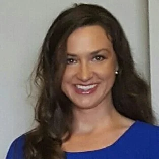 A woman with long dark hair wearing a blue top, smiling at the camera, inside a plain room.