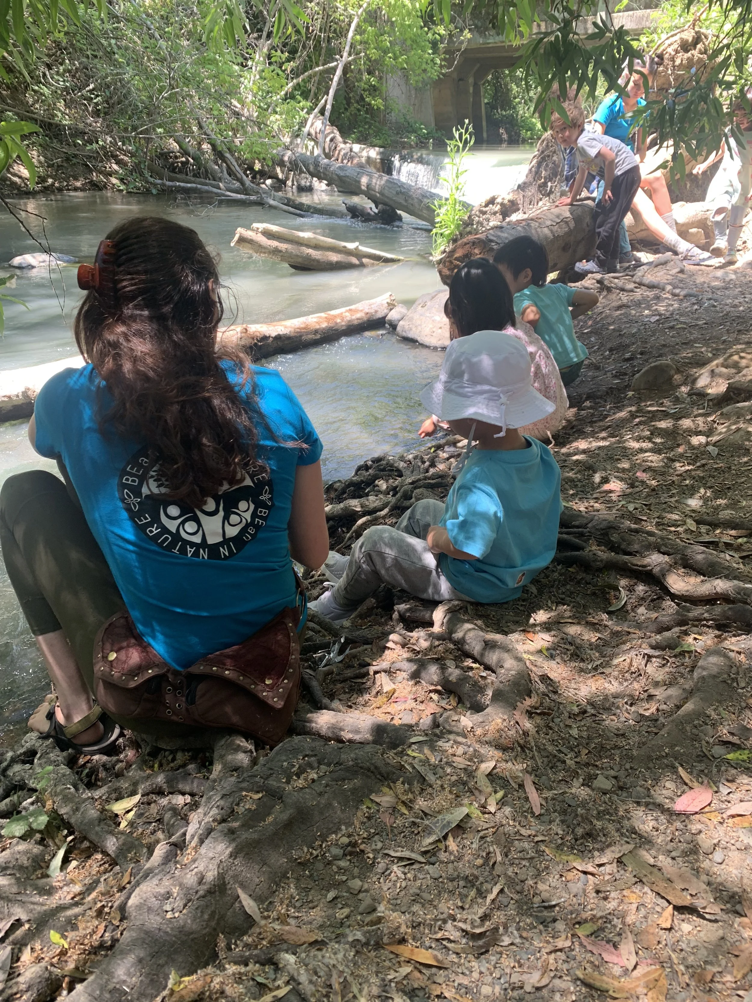 Children and a woman sitting by a riverbank with fallen trees and rocks, exploring the water surrounded by thick green foliage and a bridge in the background.