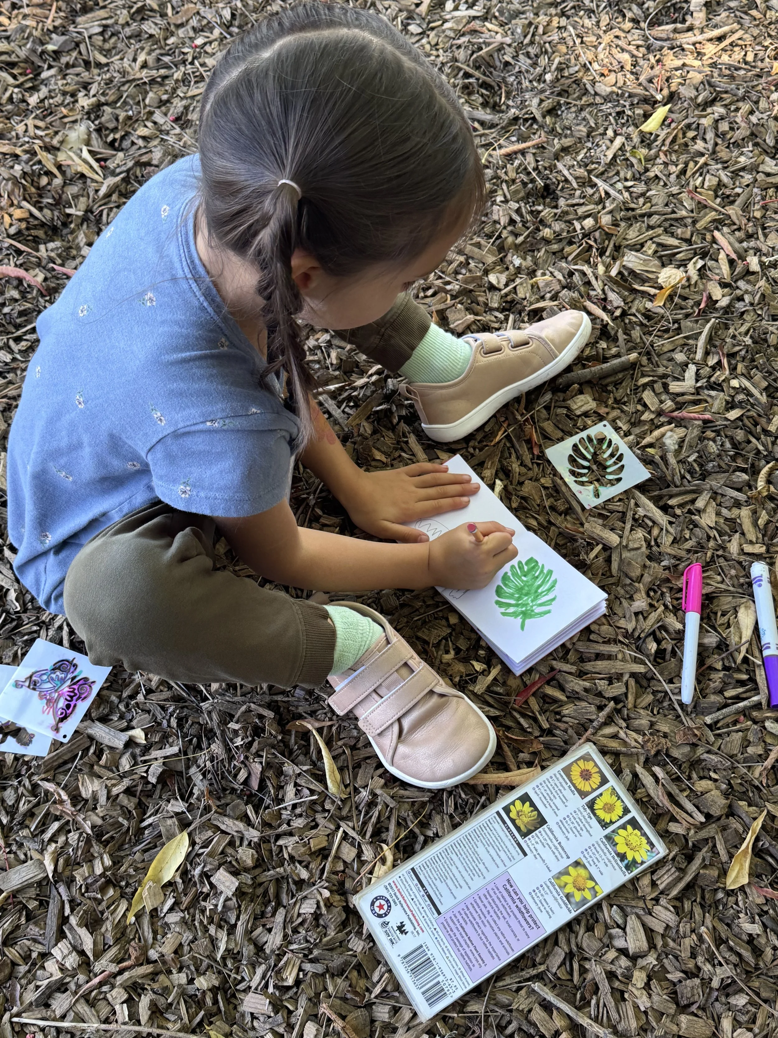 A young girl with braided hair, wearing a blue shirt and tan shoes, is sitting on the ground covered with wood chips, coloring a picture of a green leaf on a notepad. There are markers, stickers of a butterfly, a sunflower, and a leaf nearby, along w
