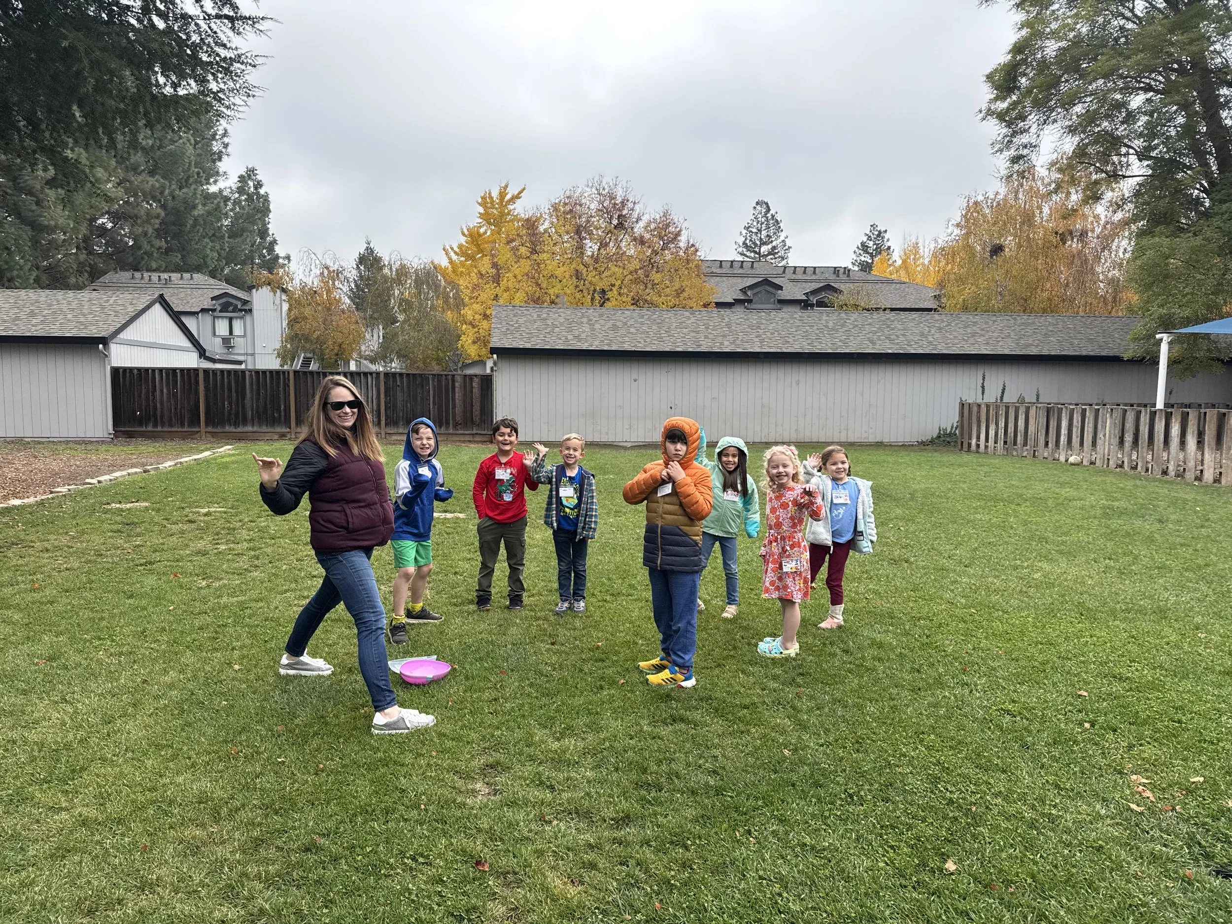 A group of children and an adult girl are playing an outdoor game in a grassy backyard, with houses and trees in the background.