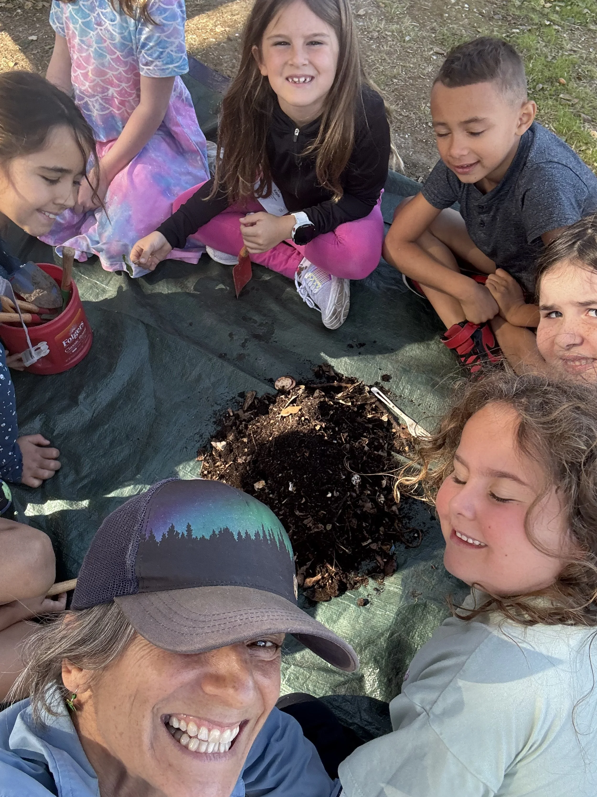 Children and an adult are gathered around a pile of soil on a tarp outdoors, smiling and looking at the camera, participating in an outdoor nature activity.
