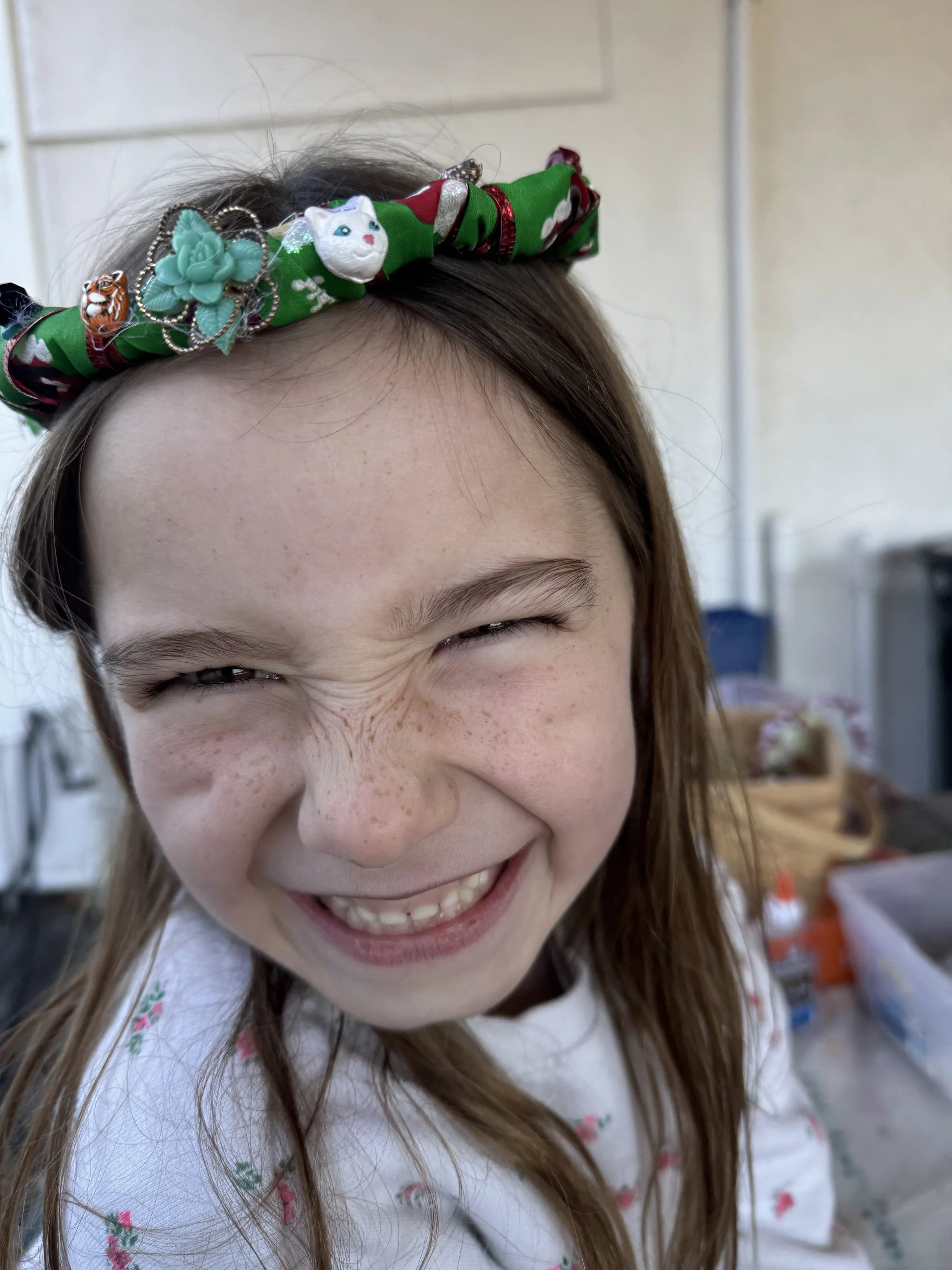 A young girl with long brown hair, squinting and smiling with her eyes closed, wearing a colorful headband decorated with charms, animals, and flowers.