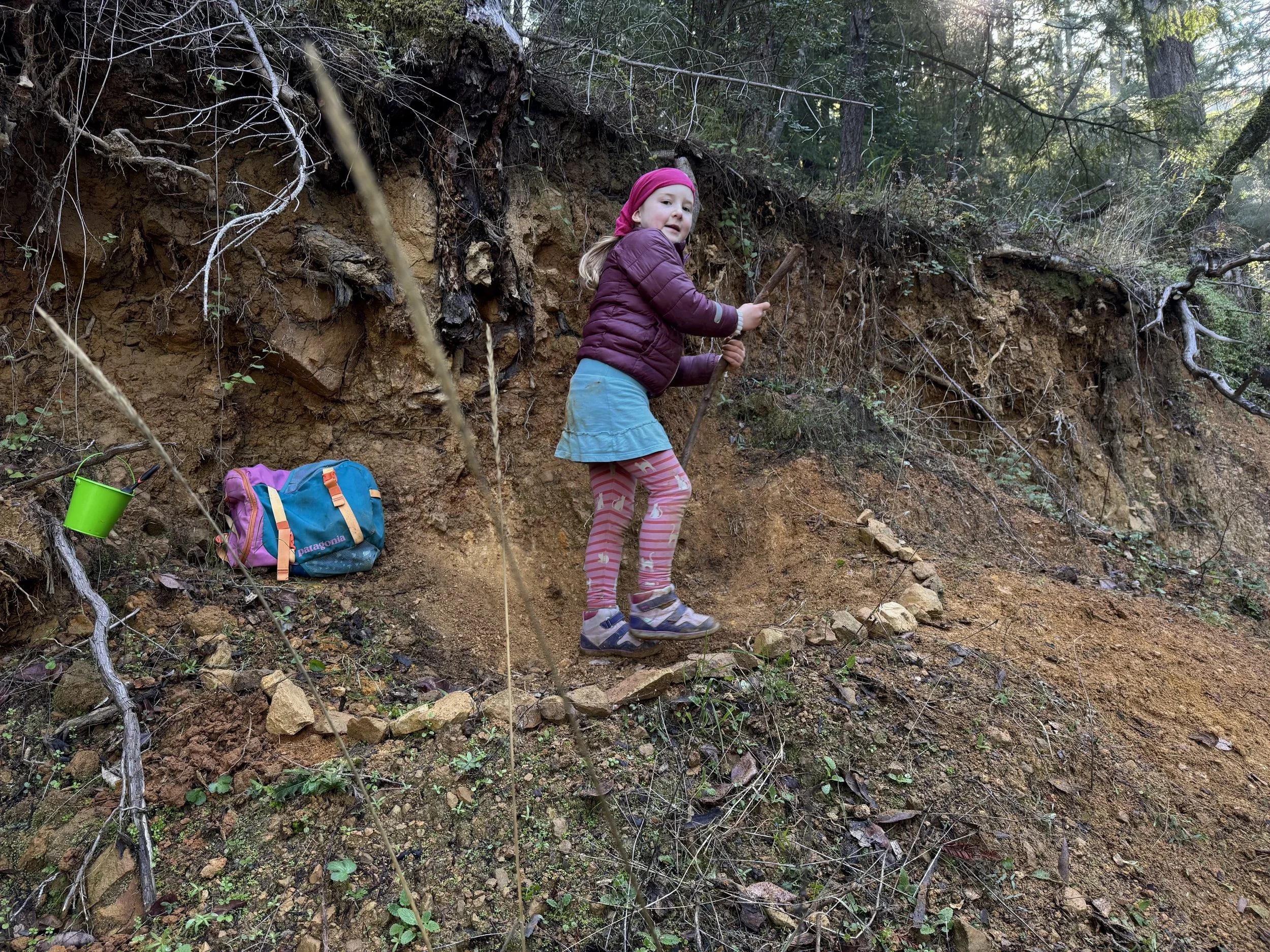 A young girl with a pink headscarf, purple jacket, pink and white striped leggings, and blue skirt, walking along a dirt trail in a forested area with trees and shrubs. There is a colorful backpack and a green cup resting on the ground nearby.