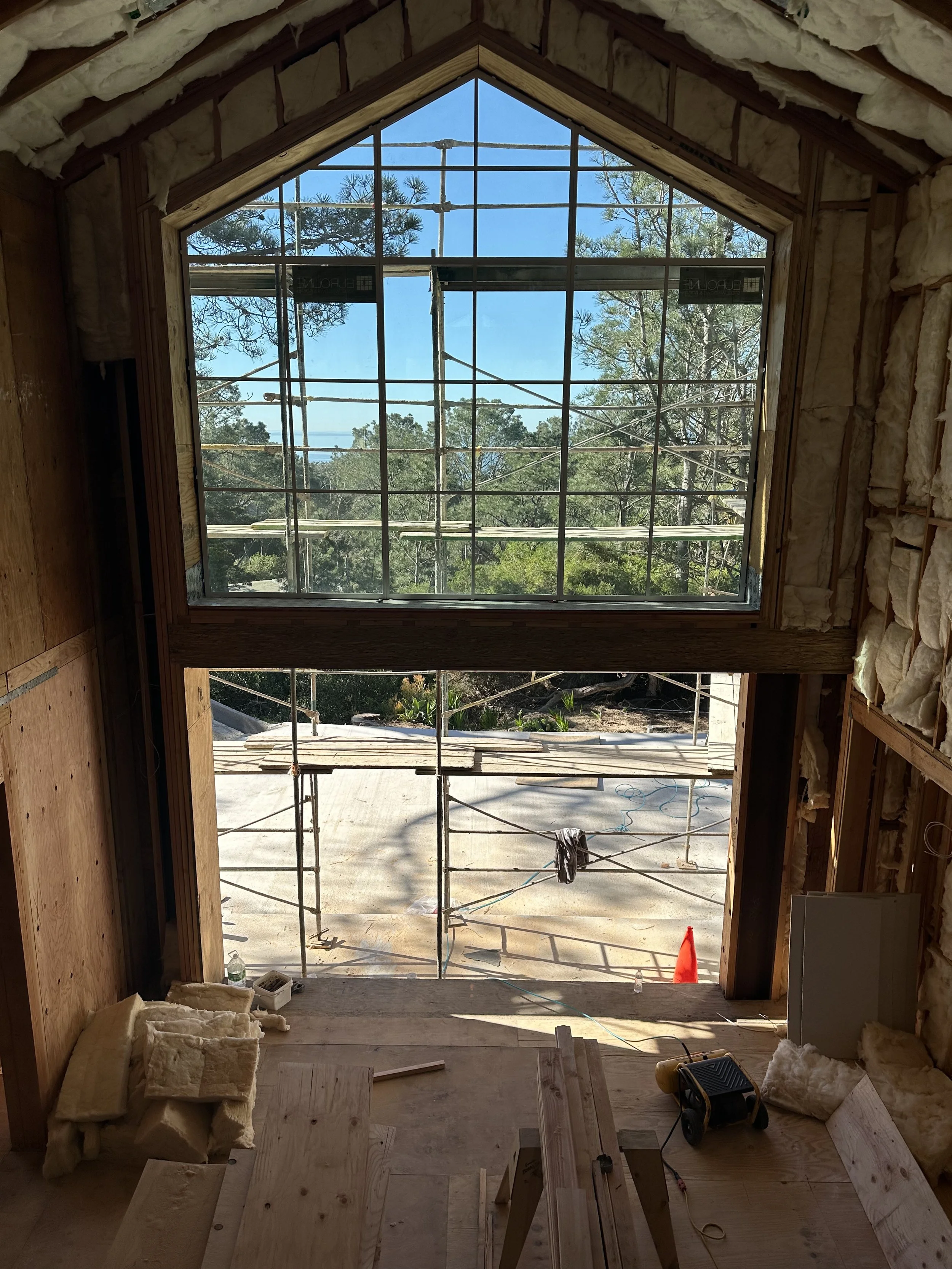 Interior of a building under construction, viewing through a large window opening with scaffolding outside, with building materials, insulation, and tools inside.
