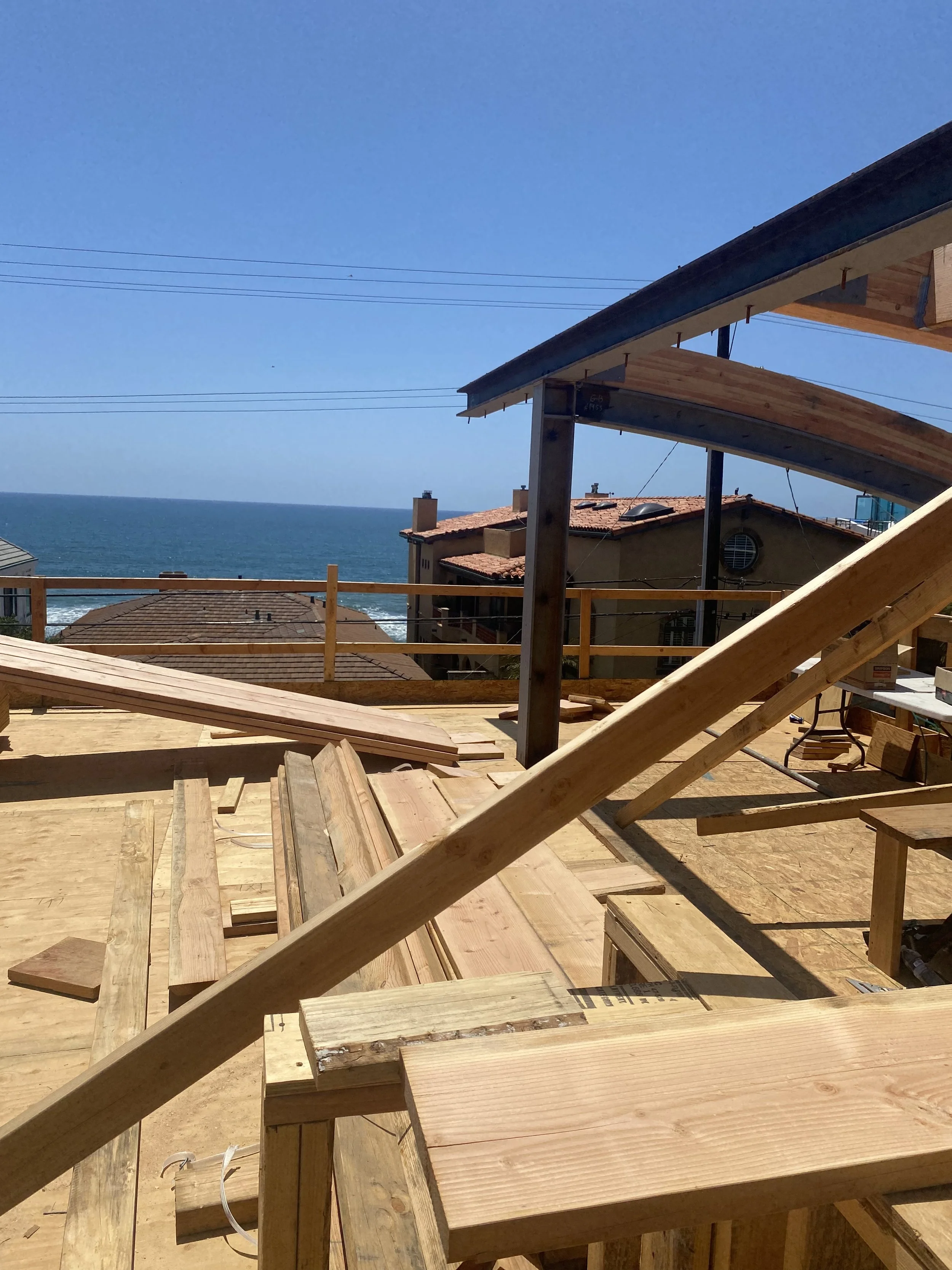 A construction site on a building's upper floor with wooden beams and framing, overlooking neighboring houses and the ocean in the distance, under a clear blue sky.
