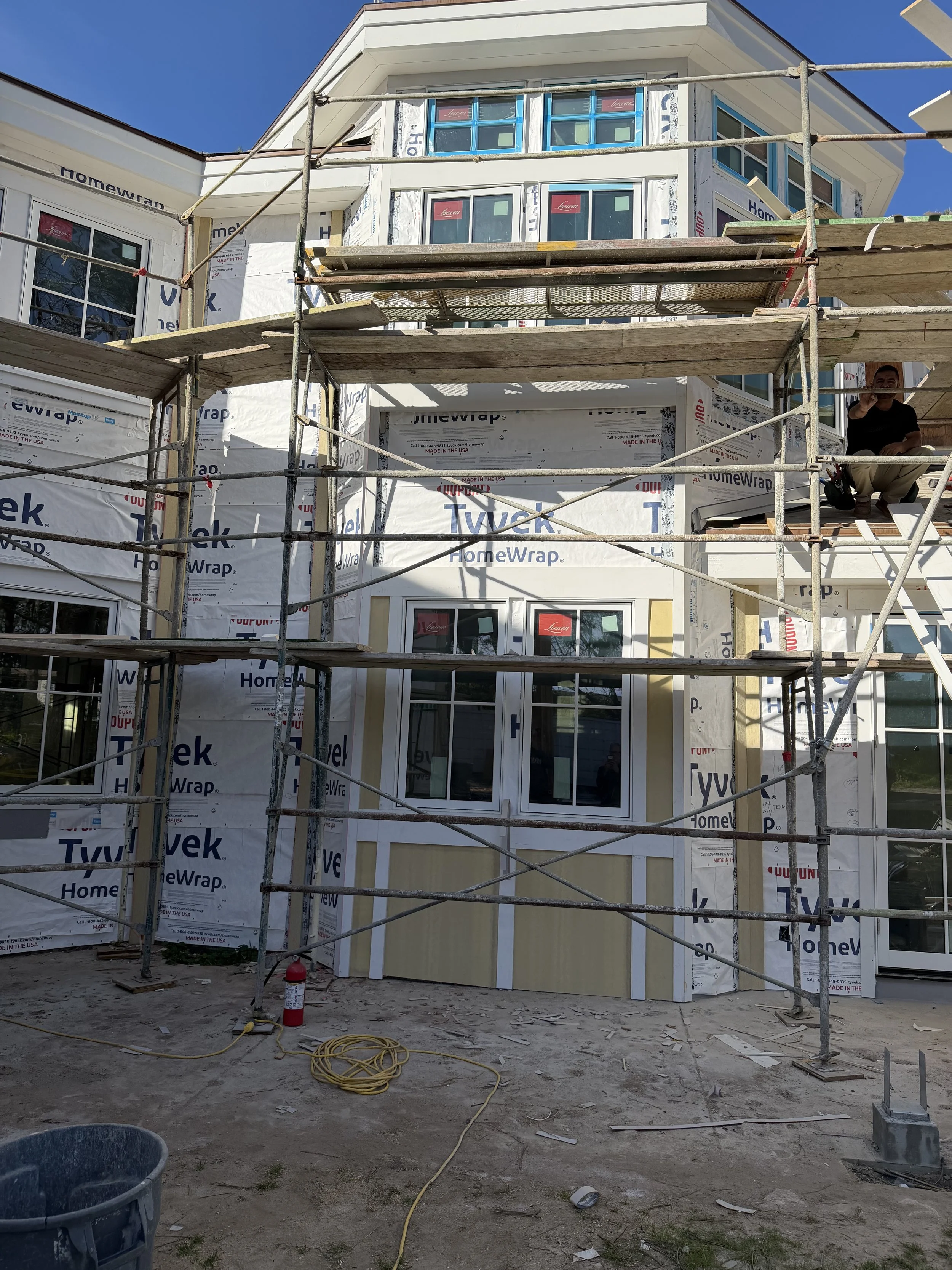 Construction site of a multi-story building with scaffolding, partially installed windows, and sheathing material. The building has a modern design with large window sections, and the site is active with construction tools and materials nearby.