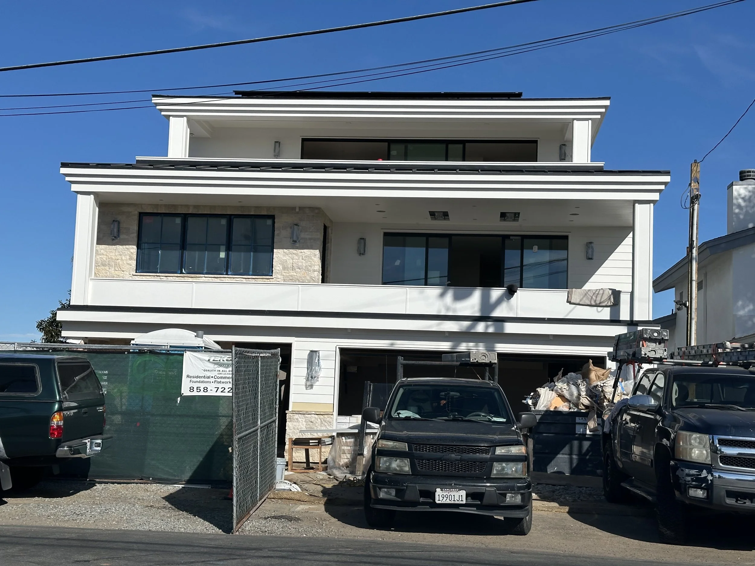 Multi-story contemporary house with large windows and a balcony, under construction or renovation, with construction materials and debris around and several vehicles parked in front, under a blue sky.