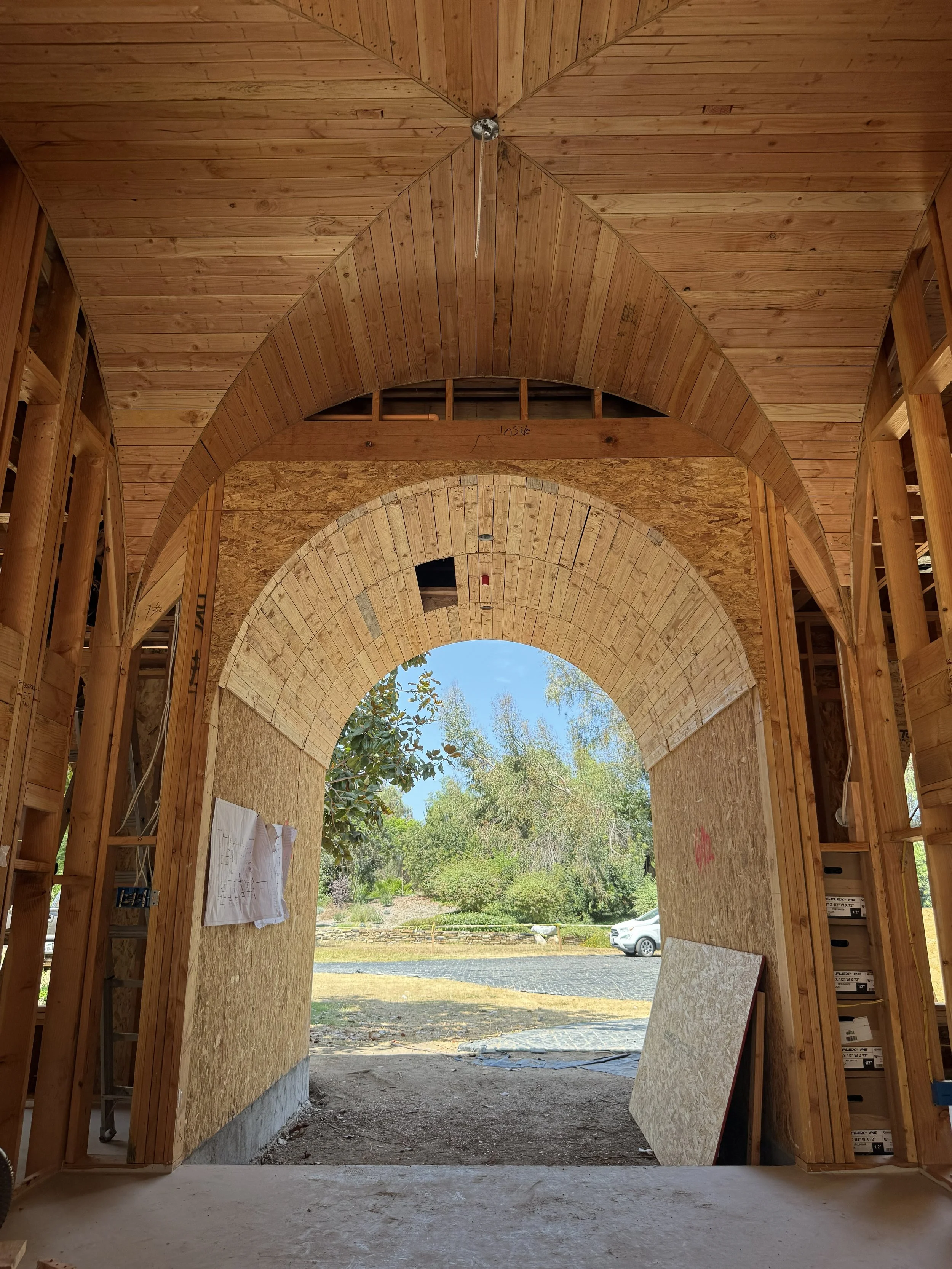 Interior view of a building under construction with wooden framing and an arched opening, showing an outdoor scene with trees, a car, and a stone wall. 
Rancho Santa Fe Covenant