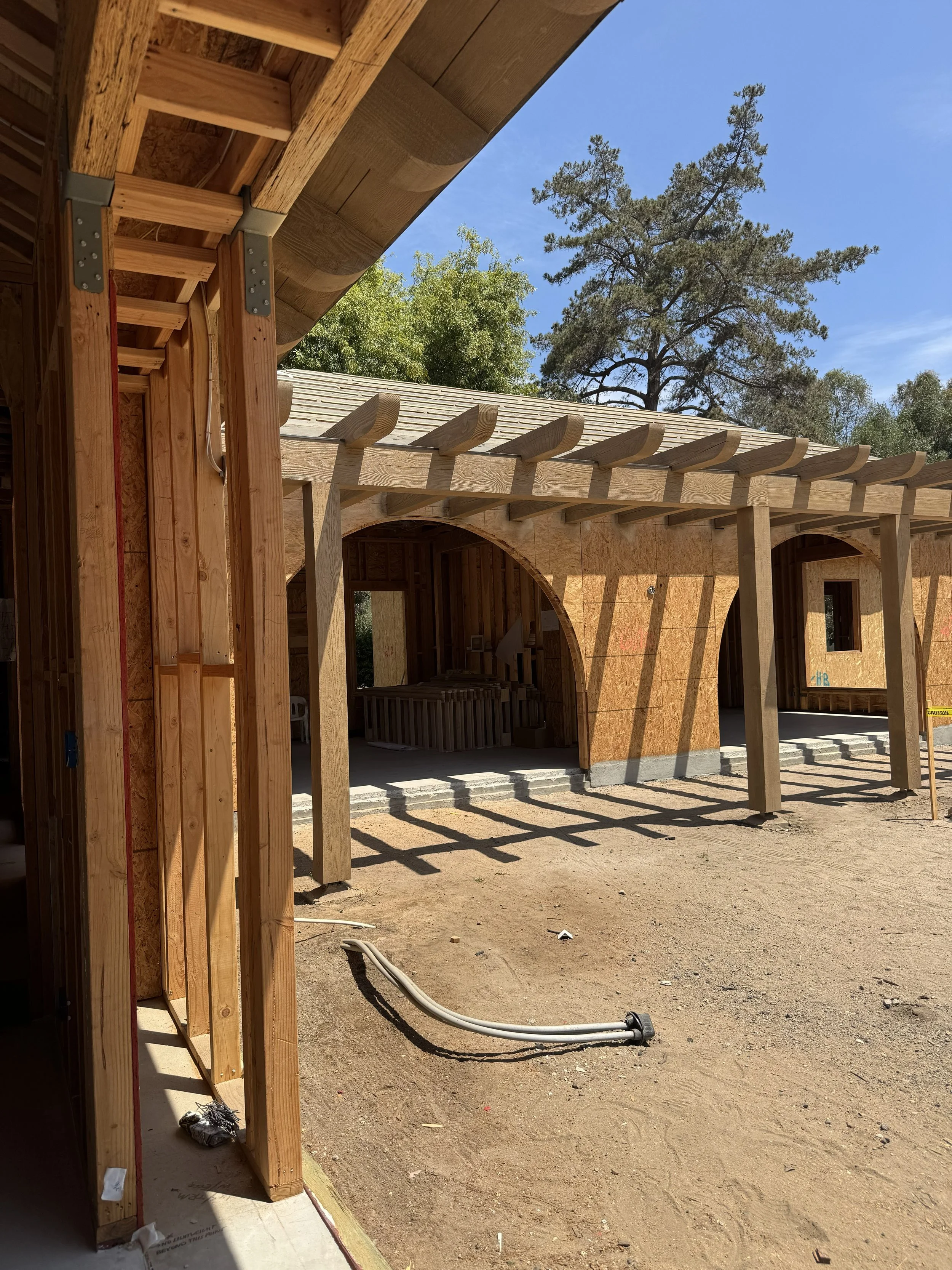 Construction site with wooden framing of a building, electrical wiring, and a dirt ground outside. Trees and a clear blue sky in the background.  Rancho Santa Fe Covenant