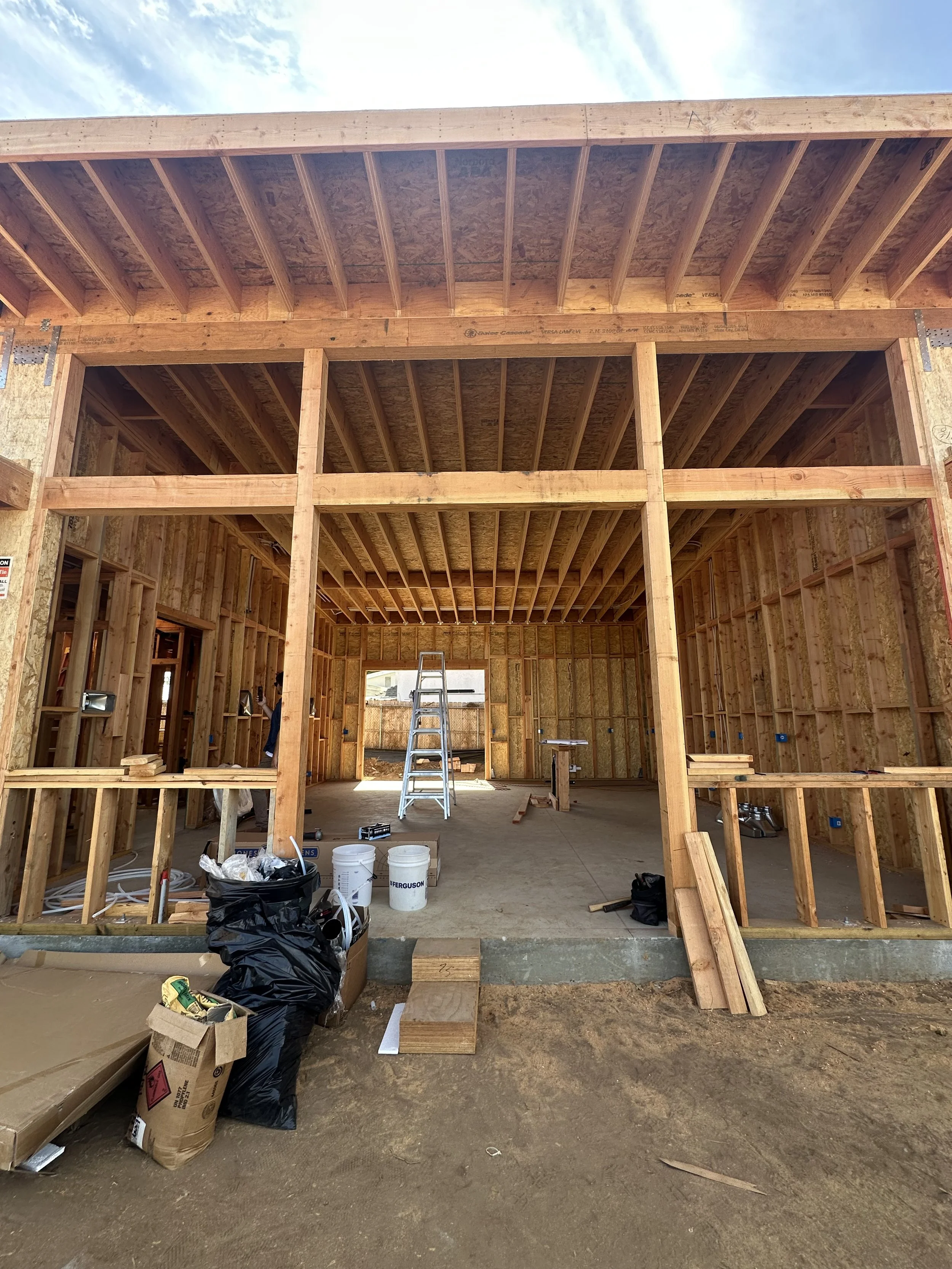 Construction site of a wooden house with framing and scaffolding, with tools, boxes, and supplies on the ground inside the structure.