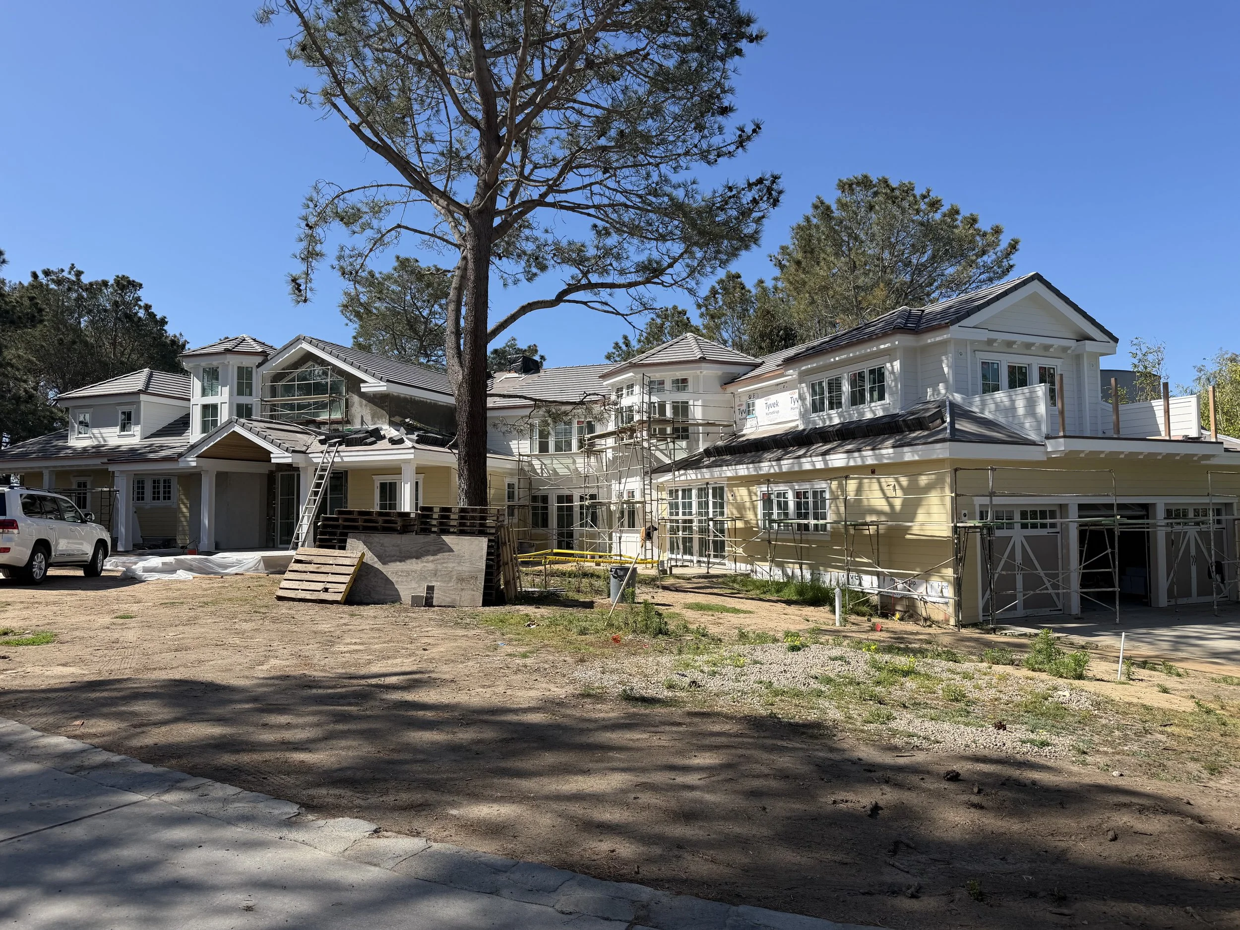 A construction site with a partially built white house, scaffolding, construction materials, a white vehicle parked in the dirt, and a large pine tree in front of the house under a clear blue sky.