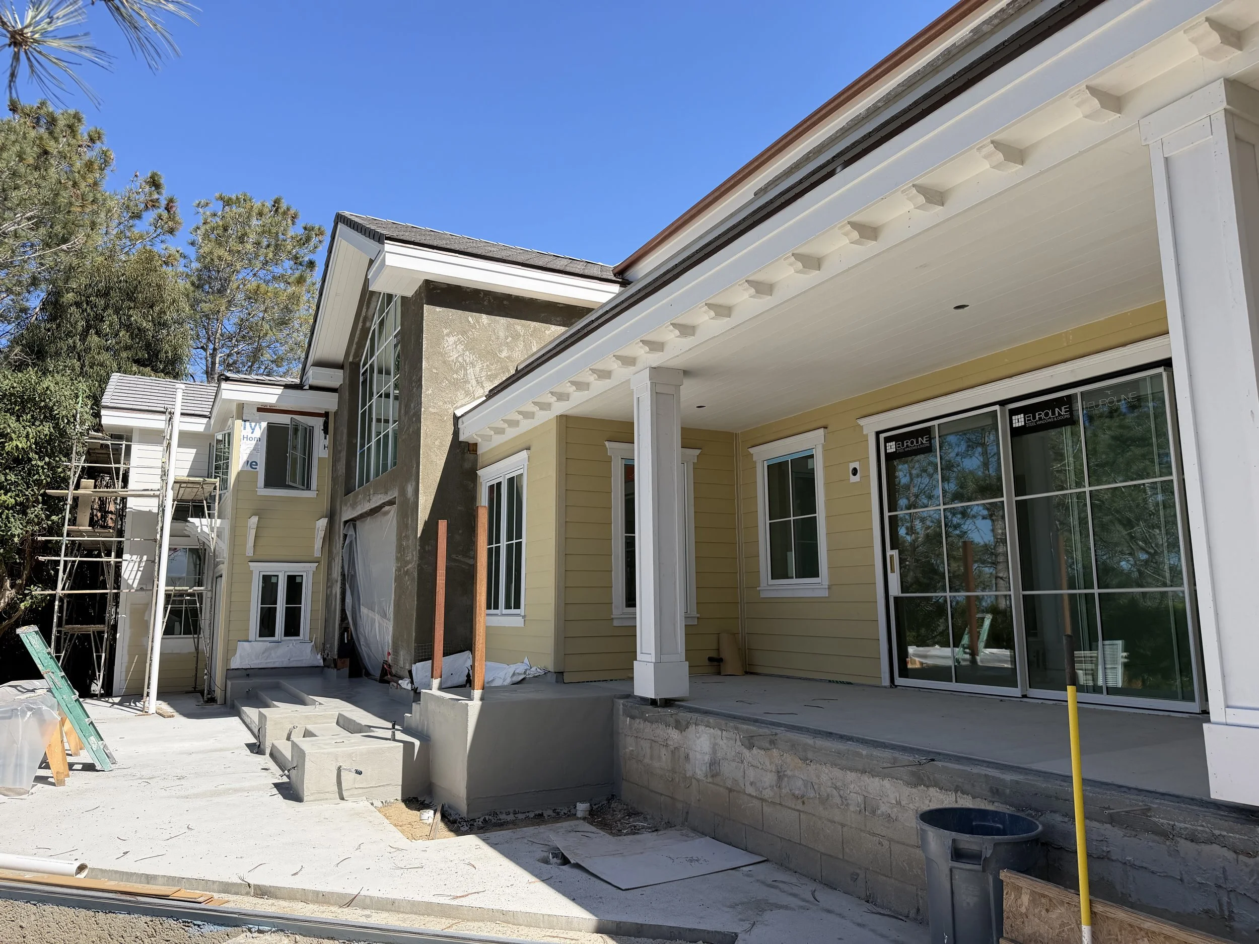 Partial view of a house under construction with yellow siding, white trim, large windows, and a screened porch. Scaffolding is visible on the left side, and construction materials are scattered in the foreground. Trees are in the background under a c