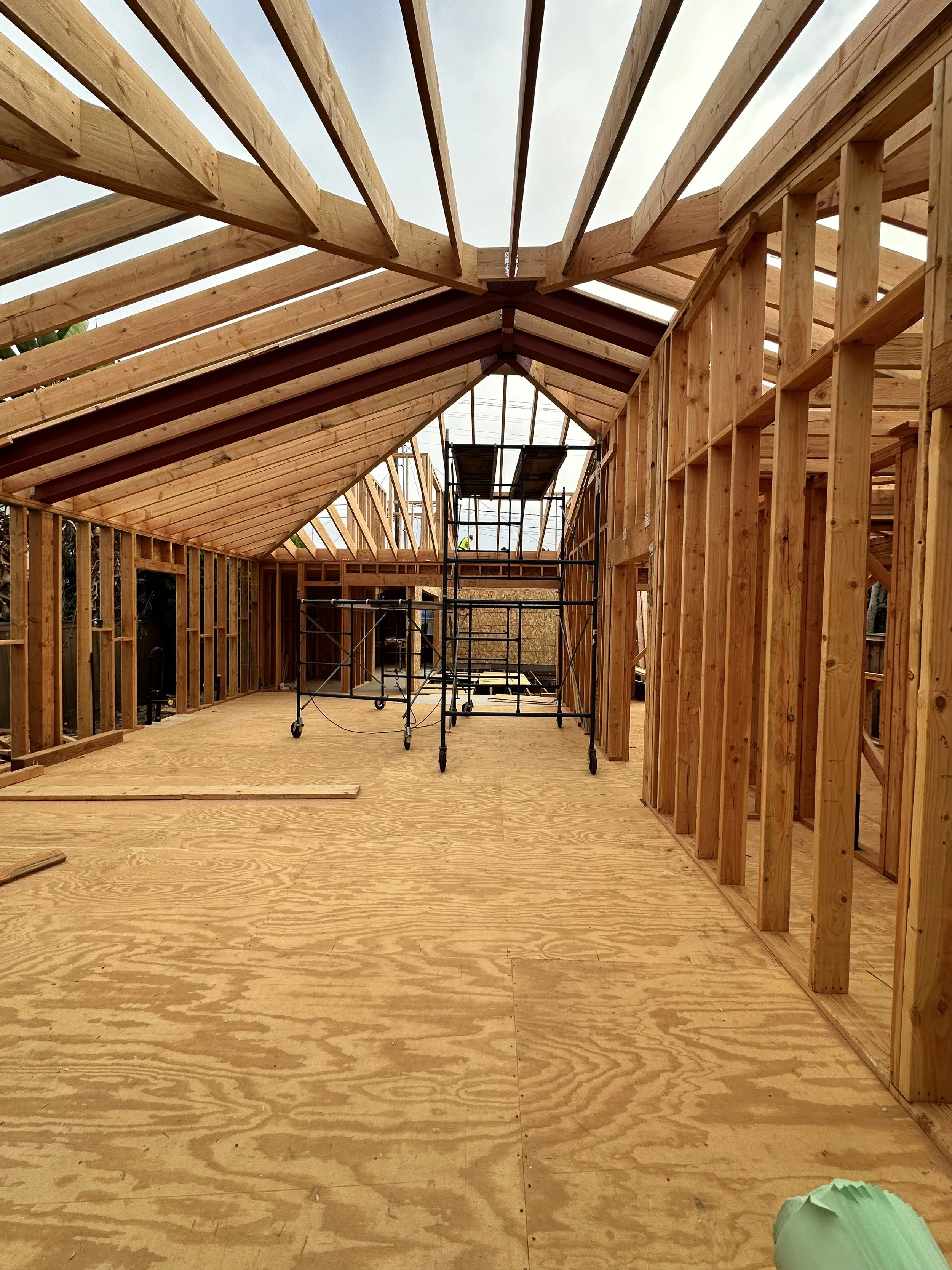 Wooden framing of a building under construction, with exposed roof trusses, scaffolding, and a plywood floor.