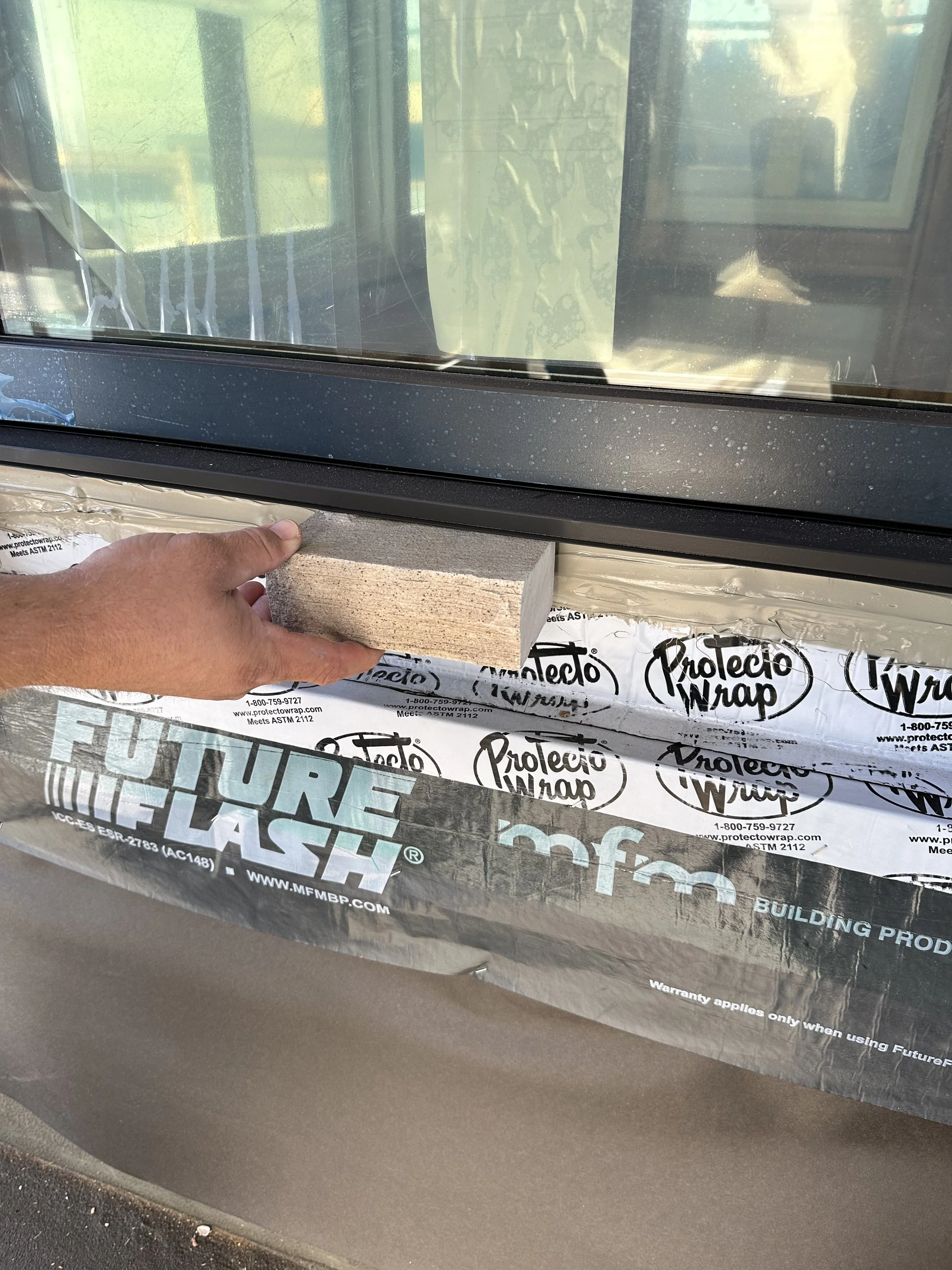 A person installing or inspecting a window with a piece of brick and a building wrap labeled 'Protecto Wrap' underneath.