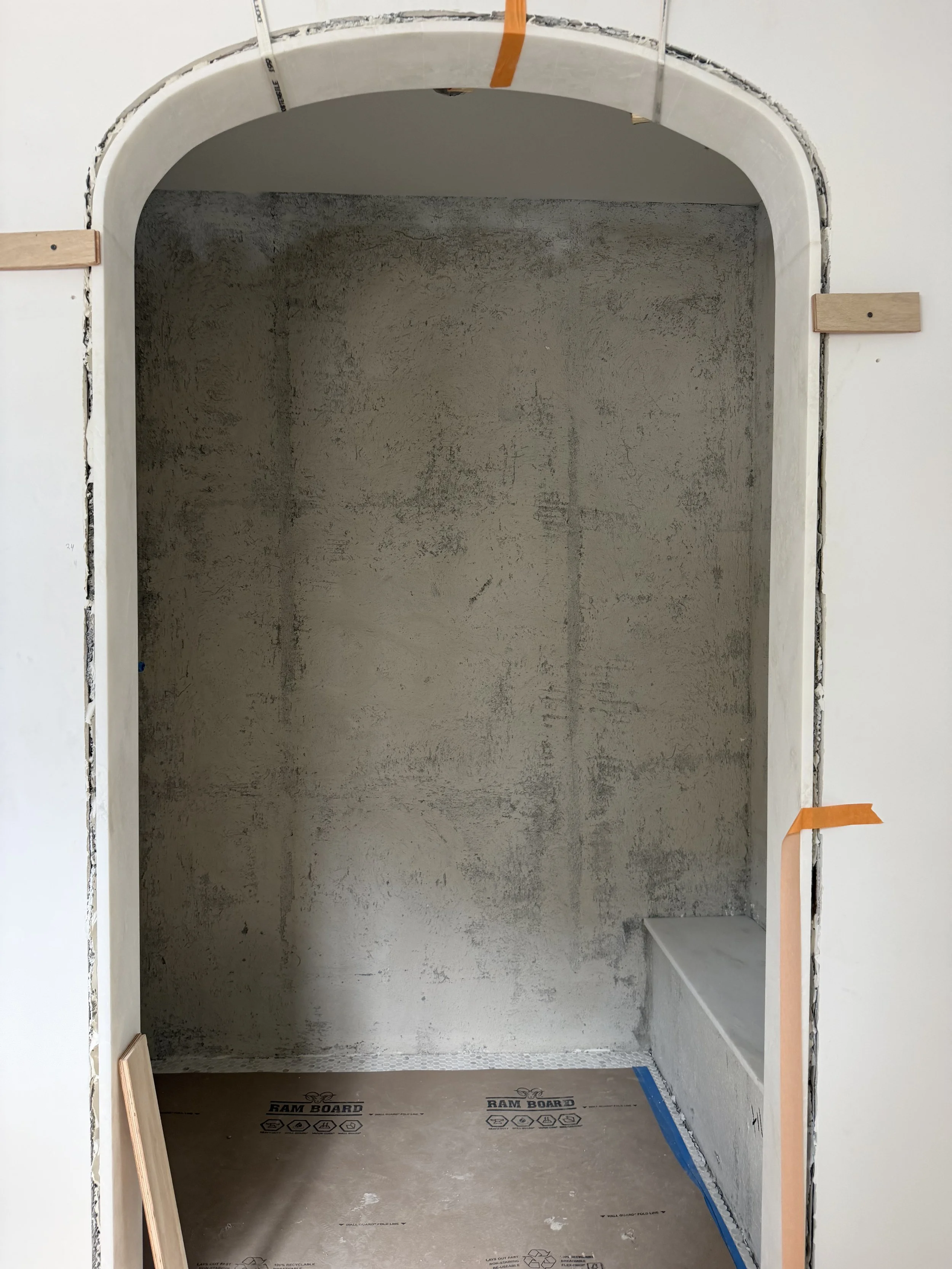 An unfinished interior space under construction with an arched opening.  There is a small wooden piece on the left side and orange tape on the right side, with other construction materials around. Del Mar