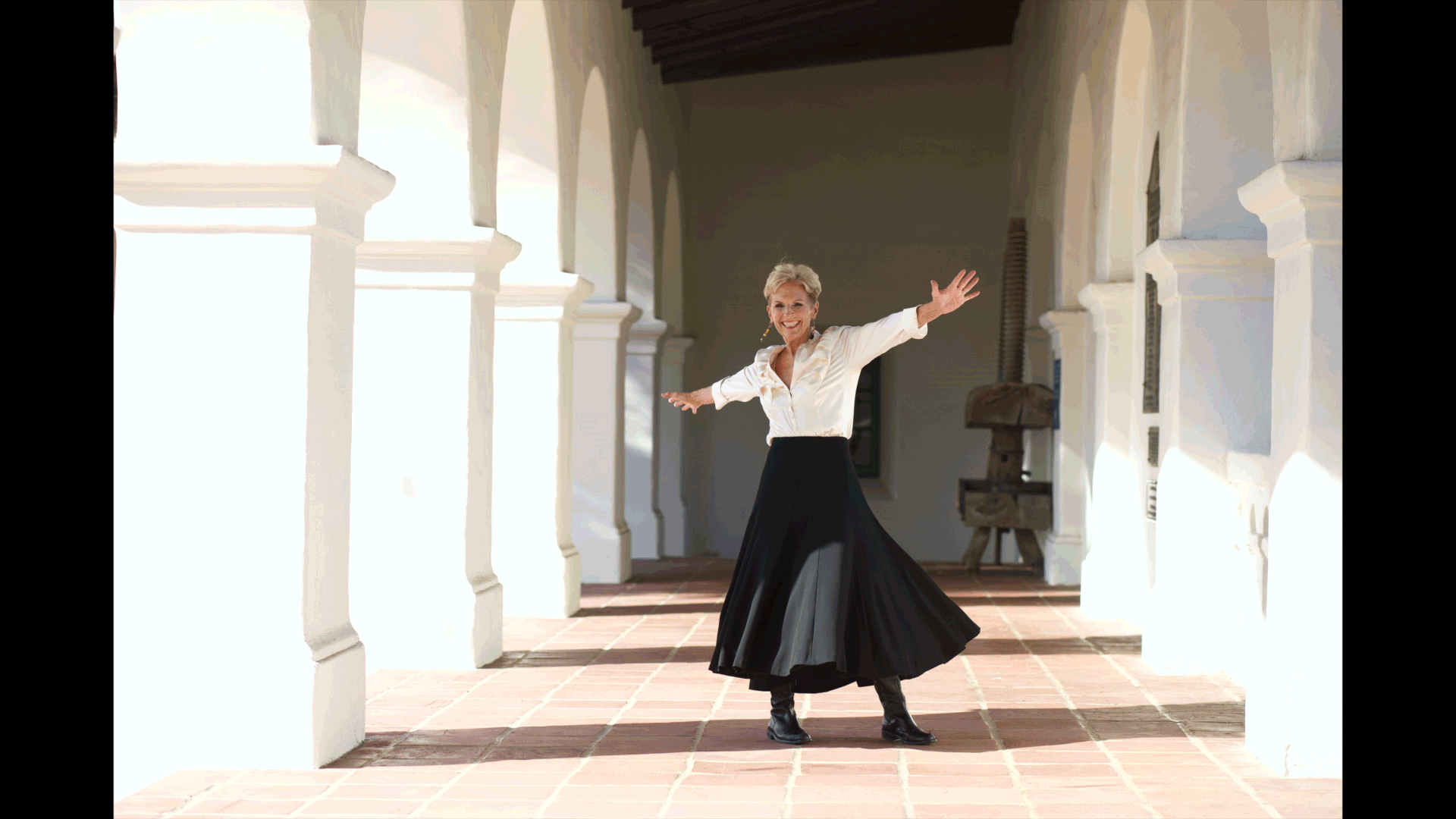 A woman with short blonde hair wearing a white blouse, black pleated skirt, and black boots, standing in a sunlit arched corridor with her arms outstretched and smiling.