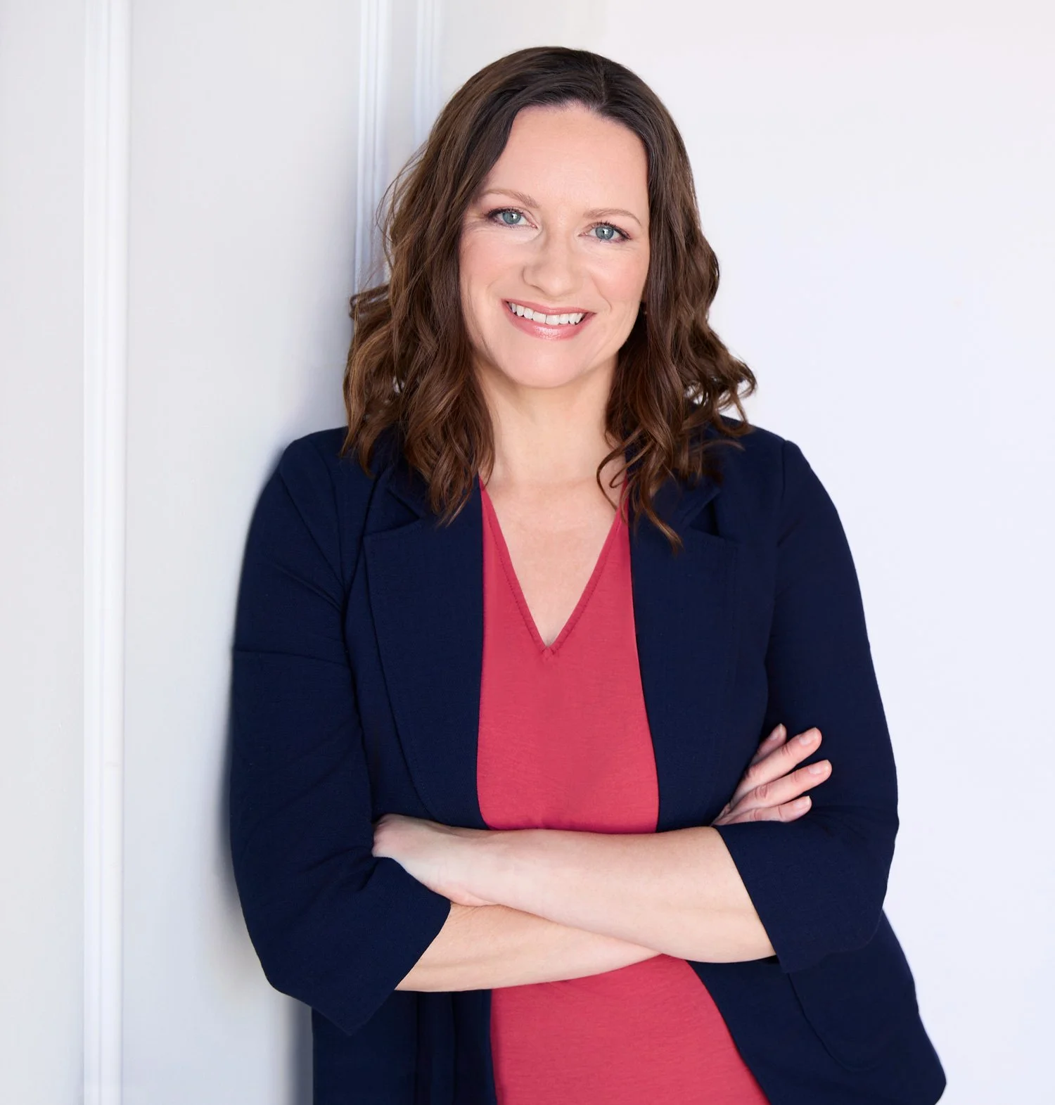 A woman with shoulder-length brown hair and blue eyes, smiling, wearing a navy blue blazer over a pink top, standing against a white wall with her arms crossed.
