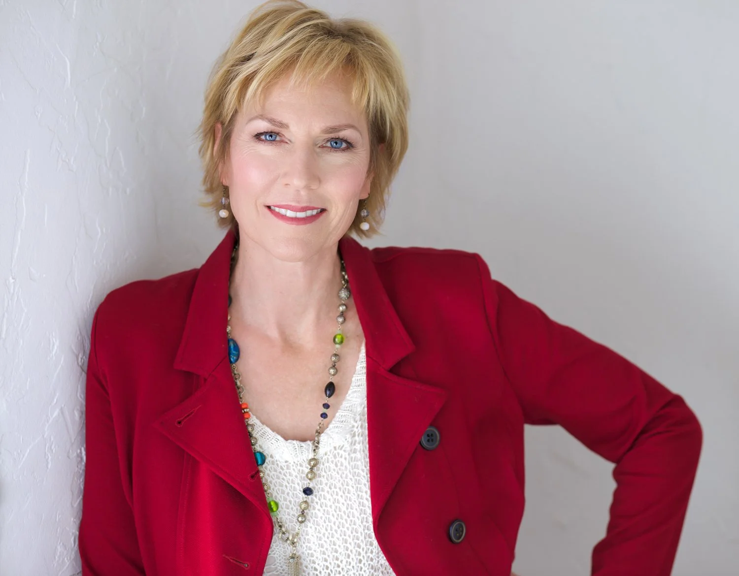A woman with short blond hair, blue eyes, and a friendly smile, dressed in a red blazer over a white lace top, standing against a white textured wall.