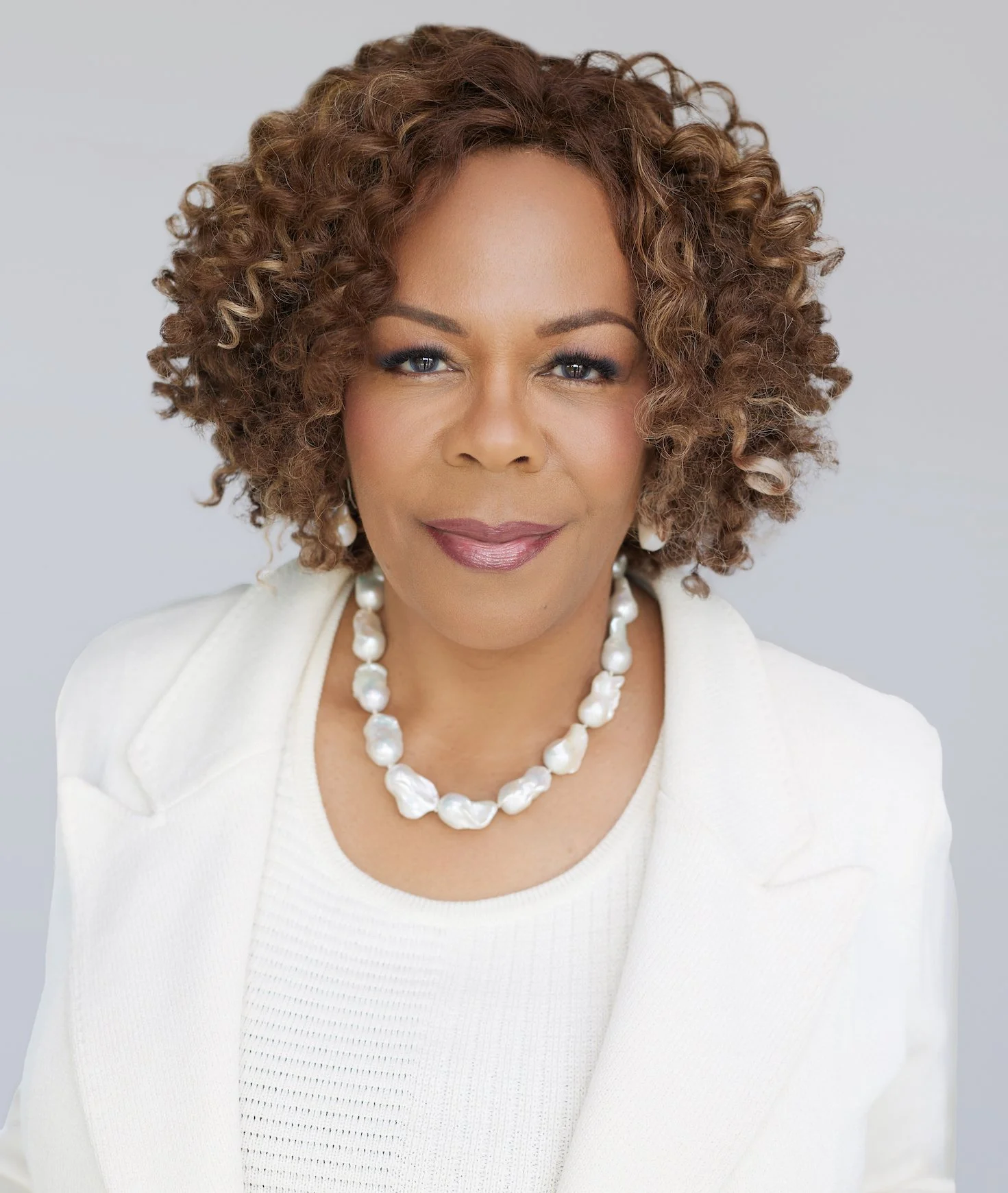 A woman with curly brown hair wearing a white blazer, white top, and a pearl necklace against a plain light gray background.