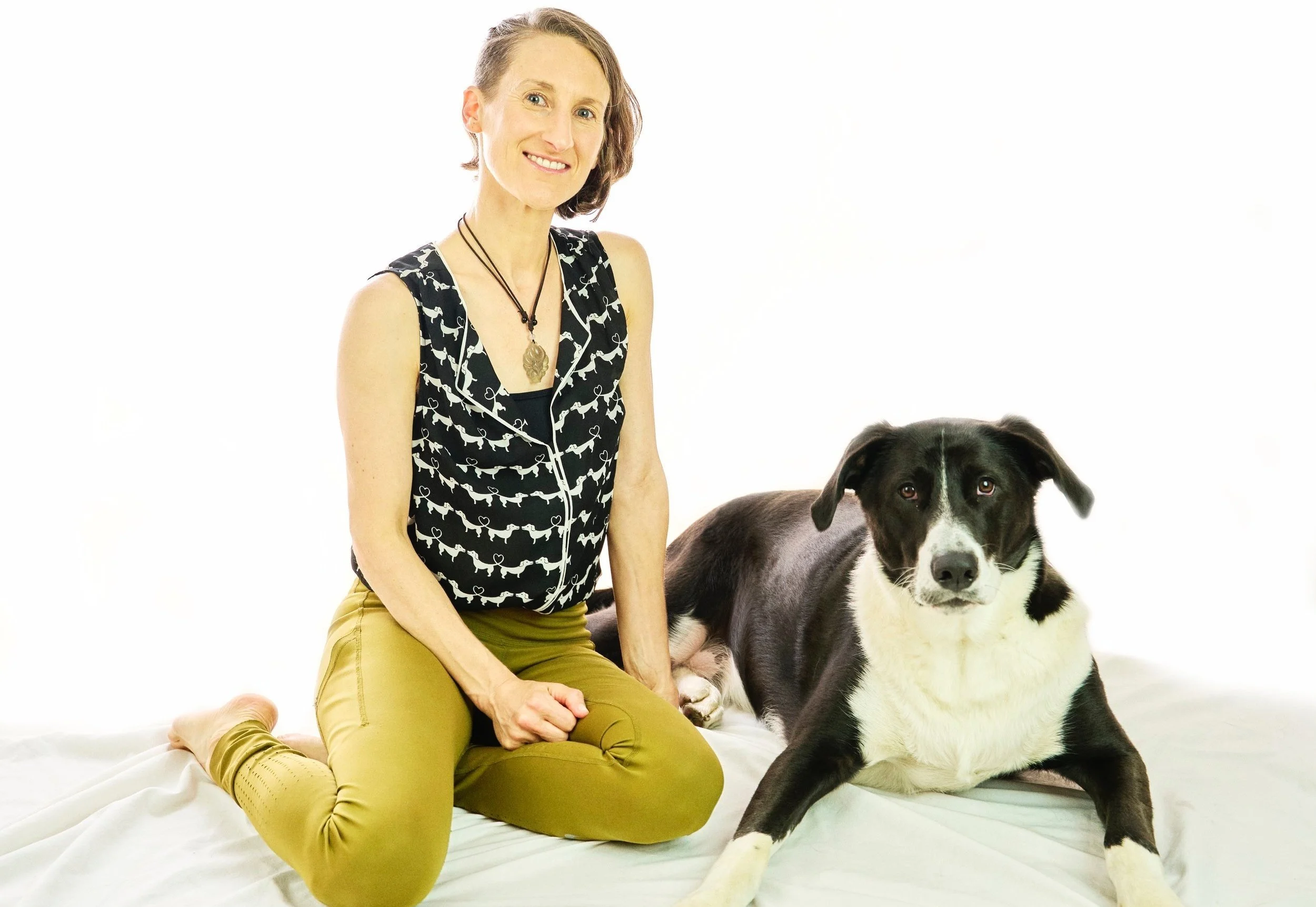 Image shows Sharon, seated on a white background with a black-and-white top and seaweed green leggings. Beside her is Muggins, a large black-and-white floppy-eared dog who captures the love of the camera.
