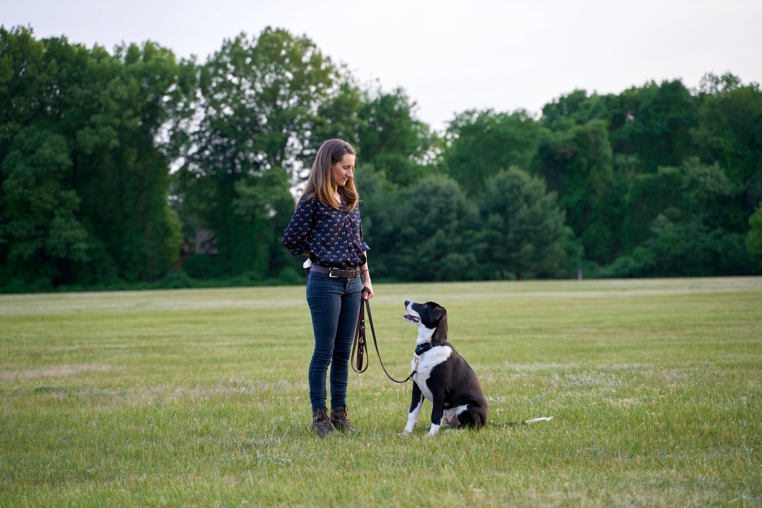 Sharon, wearing navy jeans and boots, pauses to check in with Muggins, who sits ready at her side in a grass-filled field.
